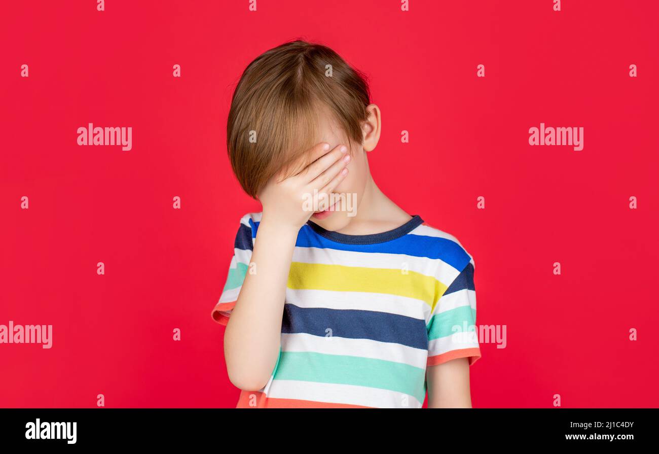 Porträt eines traurigen Jungen, der seinen Kopf mit der Hand hält, isoliert auf dem roten Hintergrund. Kleiner Junge, der Kopfschmerzen hat. Verzweiflung, Tragödie. Kopfschmerzen Kind Stockfoto