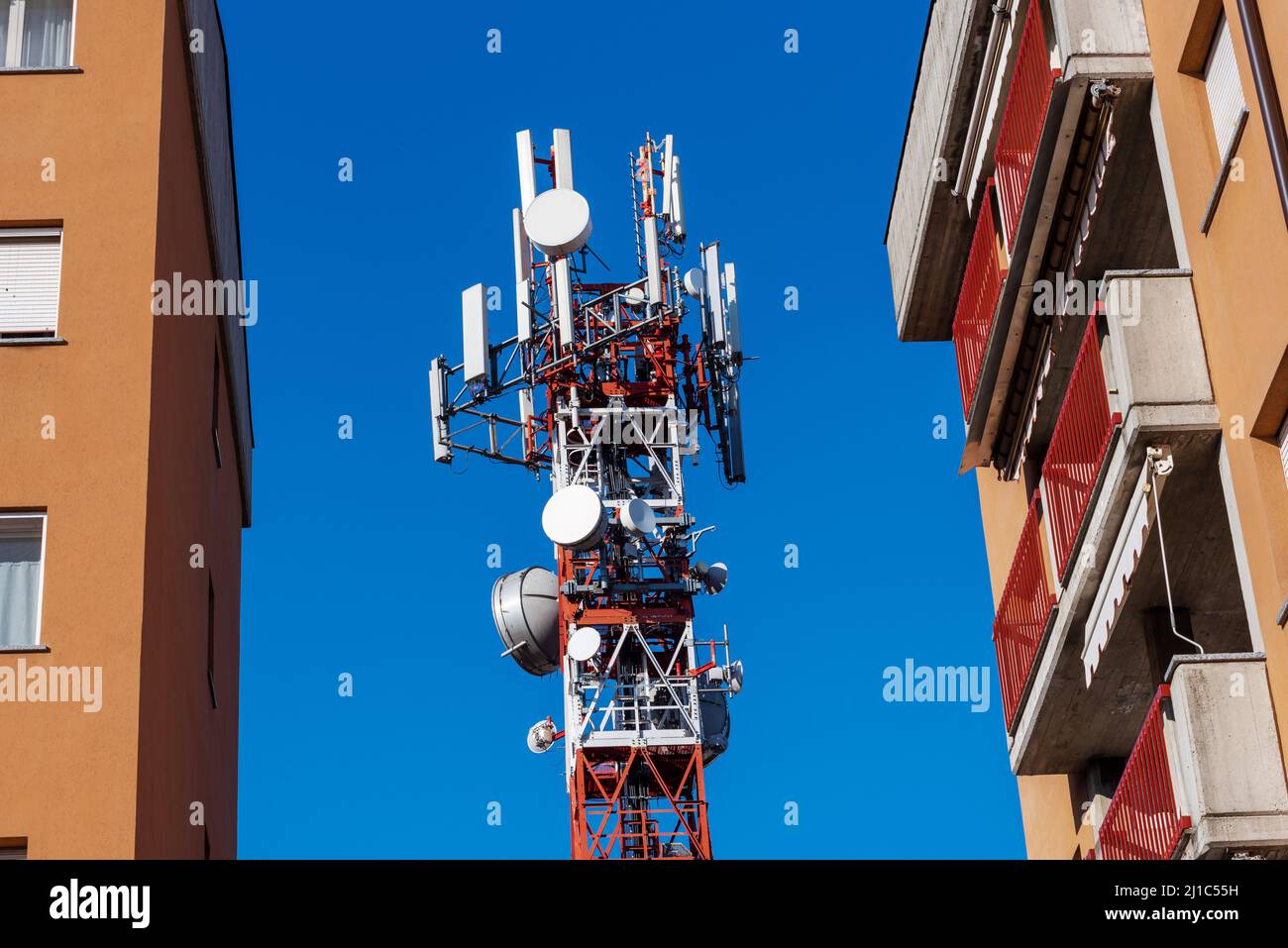 Metallturm mit Telekommunikations-Antennen und rot-weißen Gleichnissen zwischen Wohngebäuden auf blu Sky, Hochgeschwindigkeits-5G-Netzwerk und Ele Stockfoto