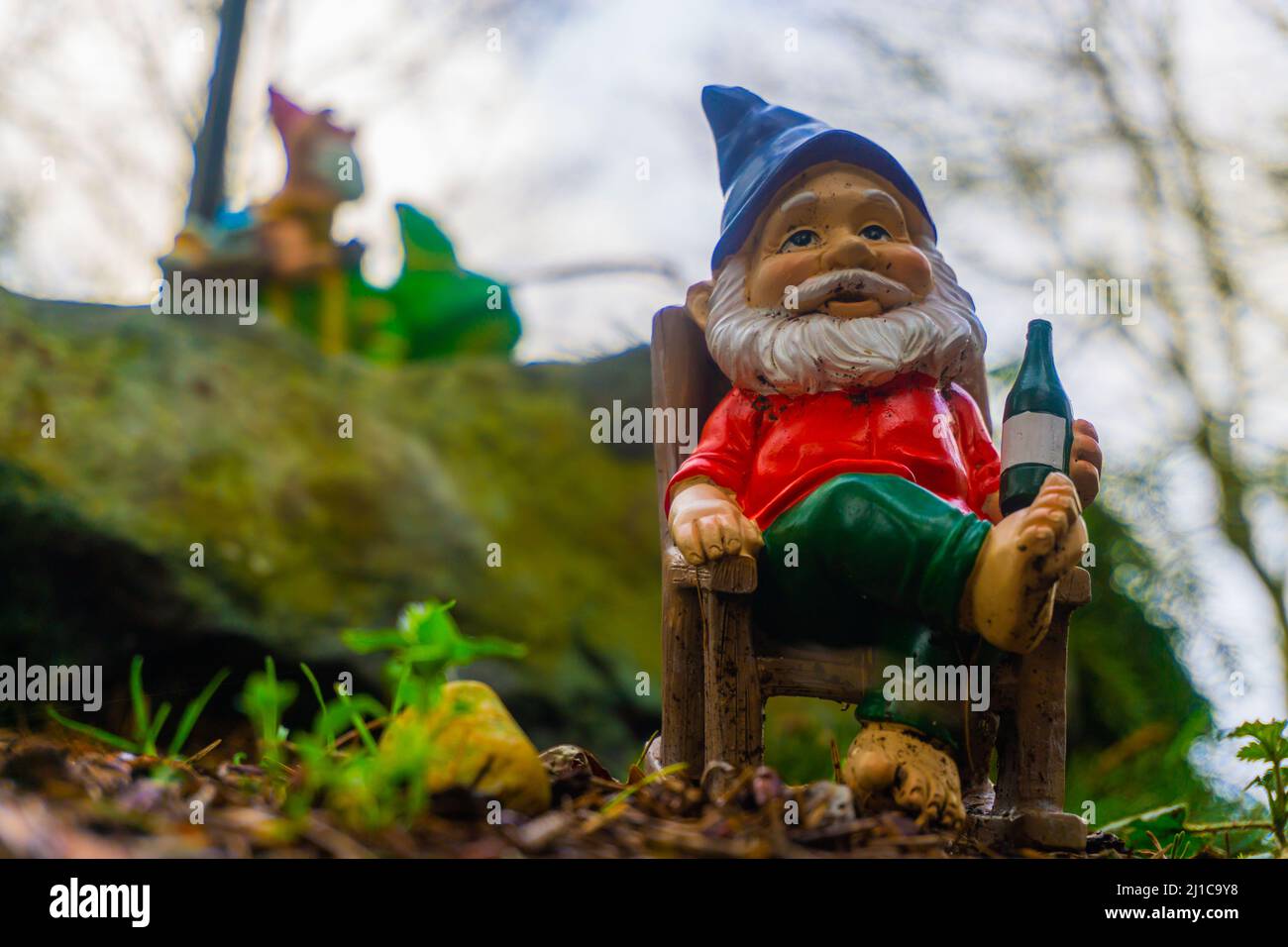 Ein Yard-Gnom entspannt sich in seinem Schaukelstuhl mit einem Gartenzwerg im Hintergrund, auf einer Eidechse sitzend. Stockfoto