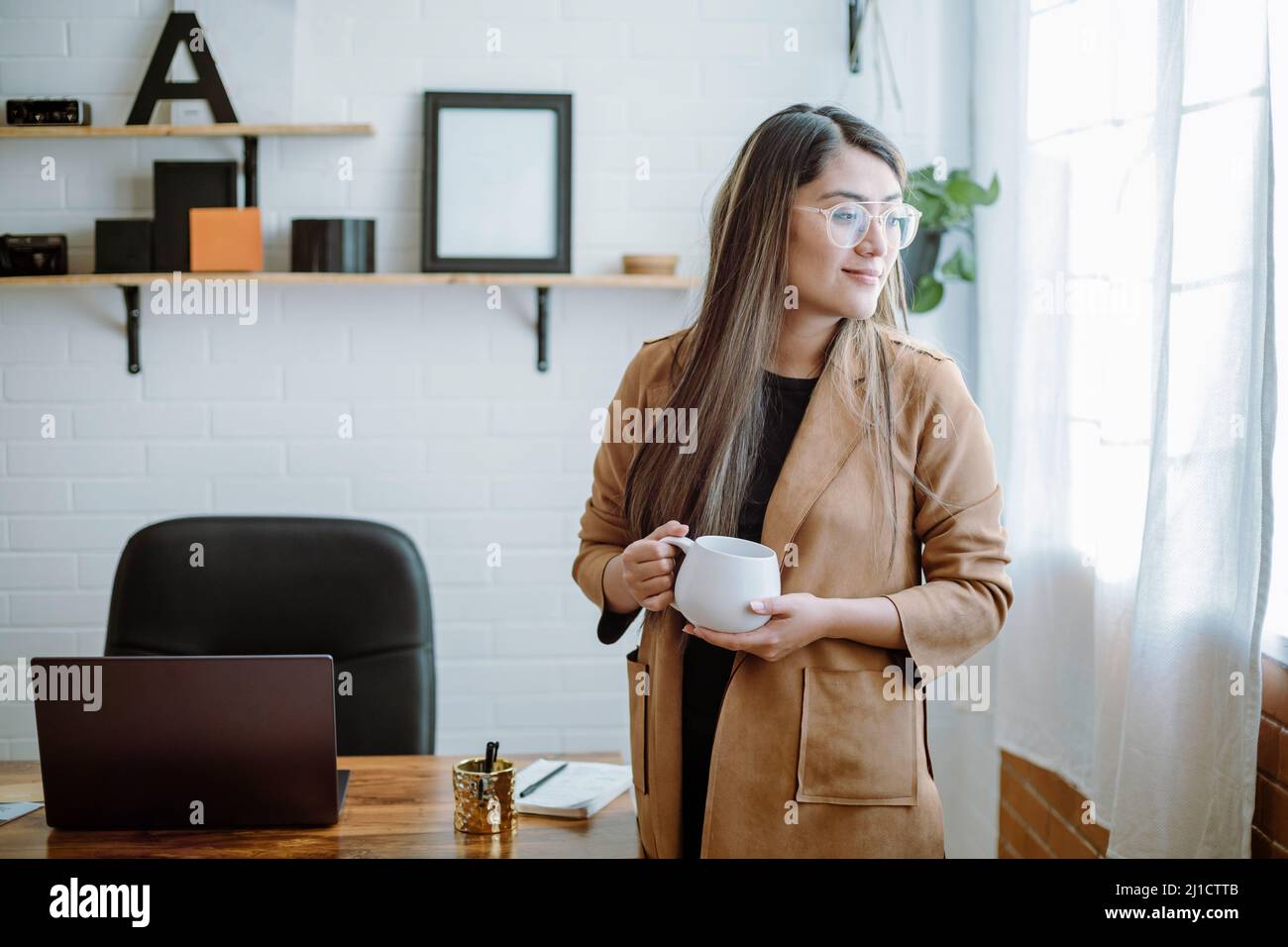 Mexikanische junge Frau trinkt aus einer weißen Tasse mit natürlicher Pose in ihrem Heimbüro Stockfoto