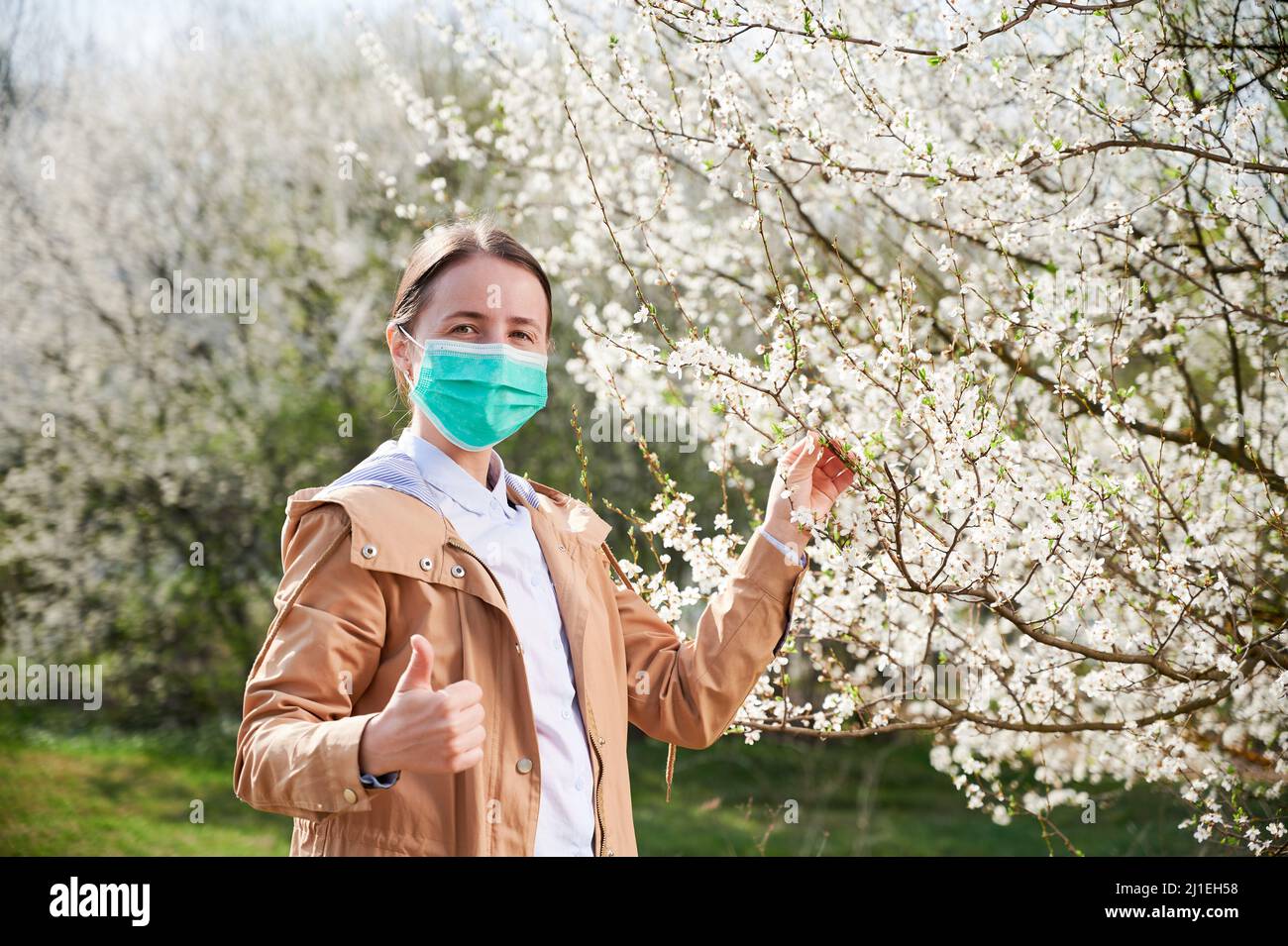 Allergische Frau, die im Frühling an einer saisonalen Allergie leidet und im Frühling im blühenden Garten posiert. Glückliche Frau, die eine medizinische Maske in der Nähe blühender Bäume trägt und die Daumen hoch zeigt. Frühjahrsallergiekonzept Stockfoto