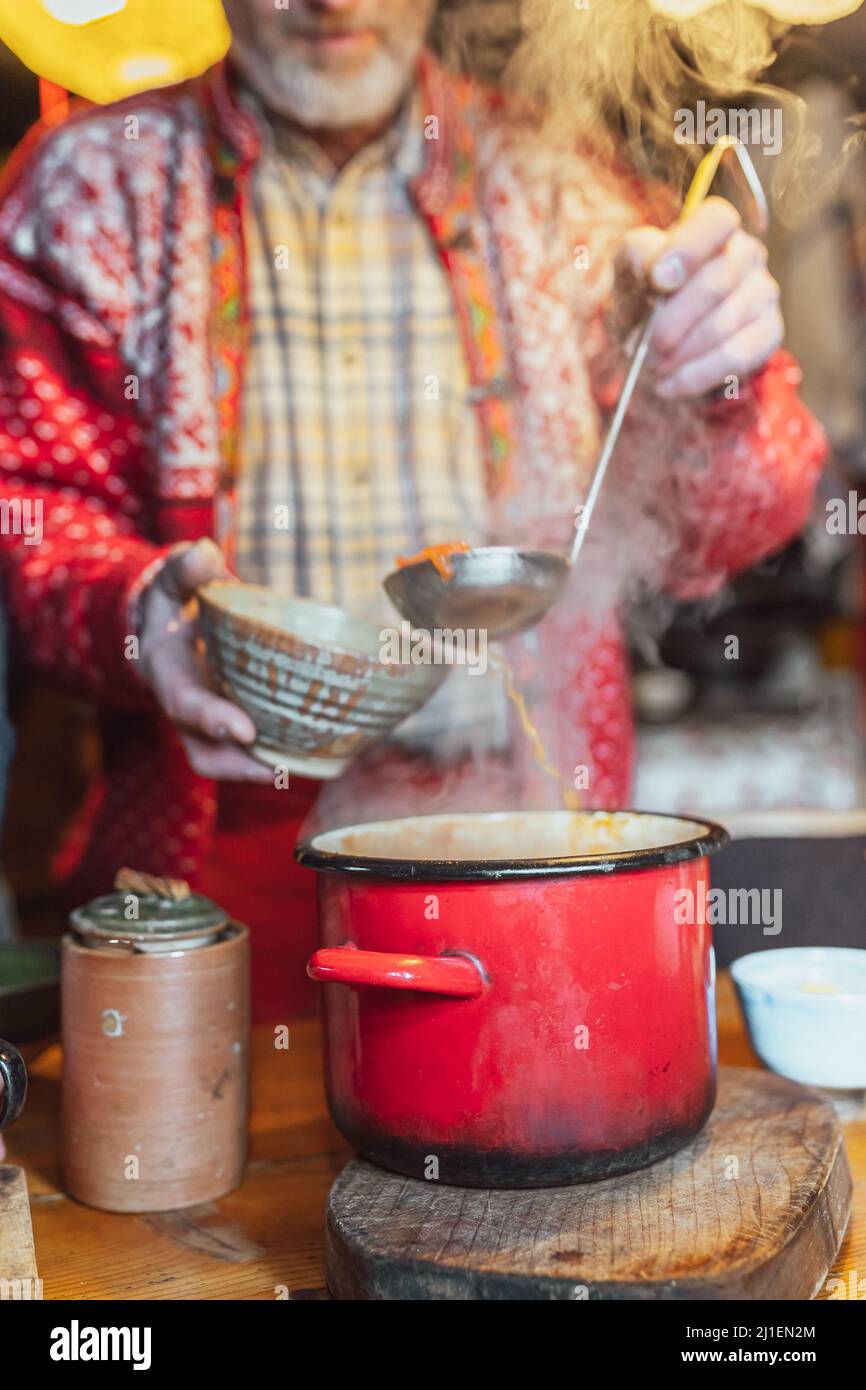 Mann in legerer rustikaler Kleidung gießt Suppe aus einem großen authentischen Topf in einen Keramikteller mit Schöpfkelle Stockfoto
