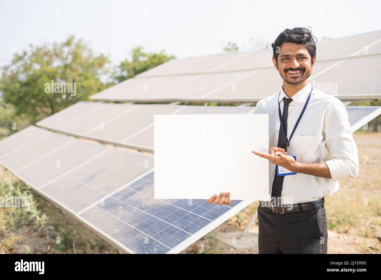 Lächelnder Bankbeamter zeigt weißes leeres Brett, indem er vor dem Solarpanel auf Ackerland die Kamera anschaut - Konzept von Botschaft, Werbung, Finanzen Stockfoto