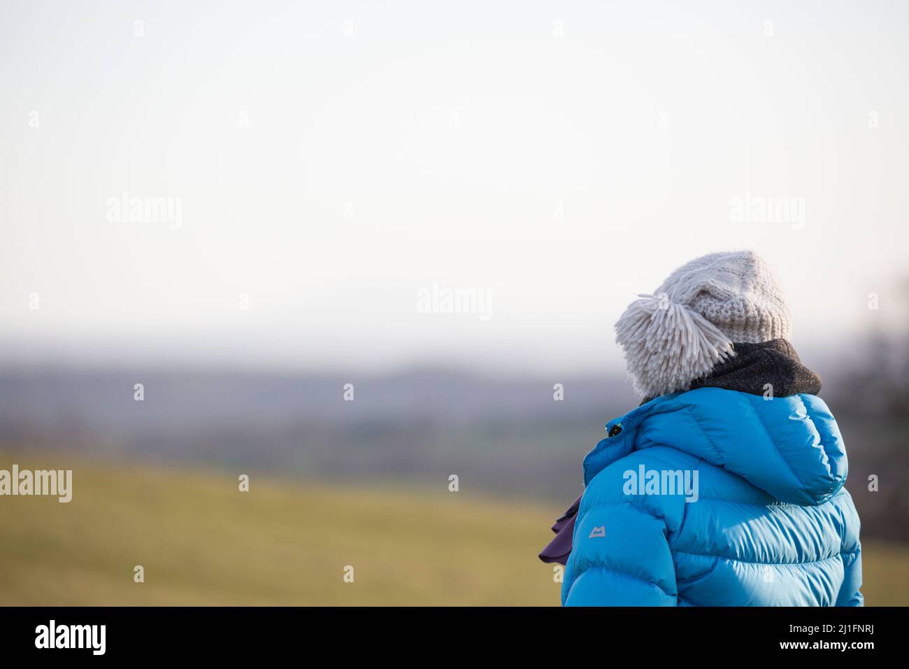Rückansicht Nahaufnahme einer isolierten Frau in wolliger Bommel-Mütze und Wintermantel im Freien auf dem ländlichen britischen Land, die in die Ferne starrt. Copyspace. Stockfoto