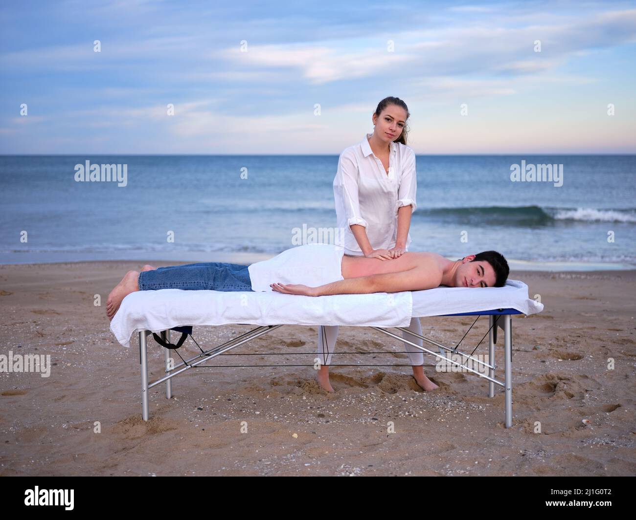 Ein junger Therapeut, der eine Chiromassage an einem jungen Mann zur Behandlung einer Lendenwirbelverletzung im Freien durchführt, und dabei auf einen Strand in Valencia blickt. Stockfoto