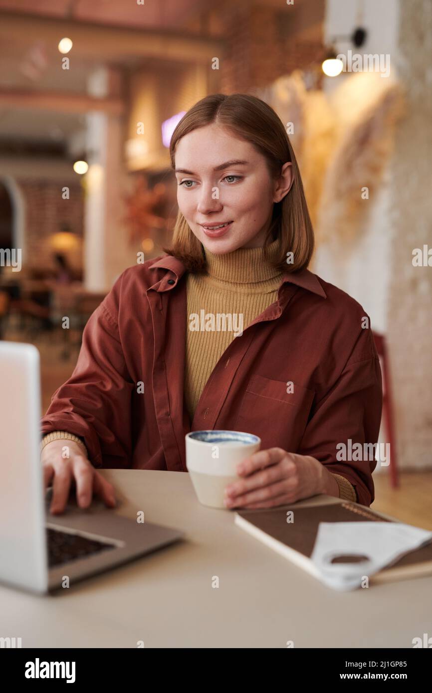 Vertikale mittlere Porträtaufnahme einer stilvollen kaukasischen Frau, die während der Kaffeepause am Kaffeetisch sitzt und einen Videoanruf auf dem Laptop führt Stockfoto