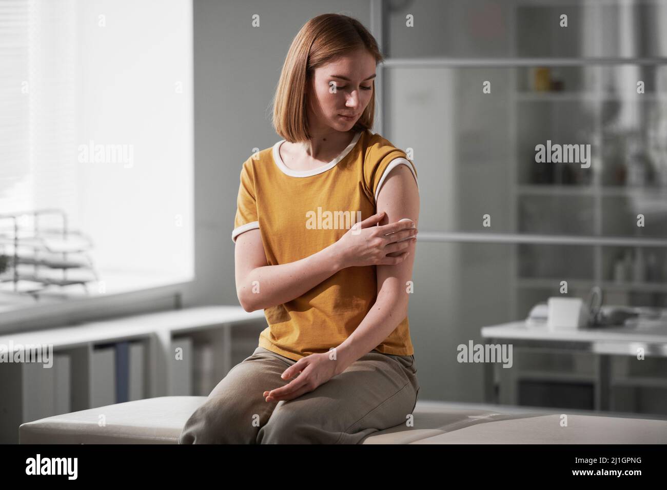 Horizontales mittellange Porträt einer jungen kaukasischen Frau, die im Impfraum sitzt und das Wattepad auf die Schulter drückt Stockfoto