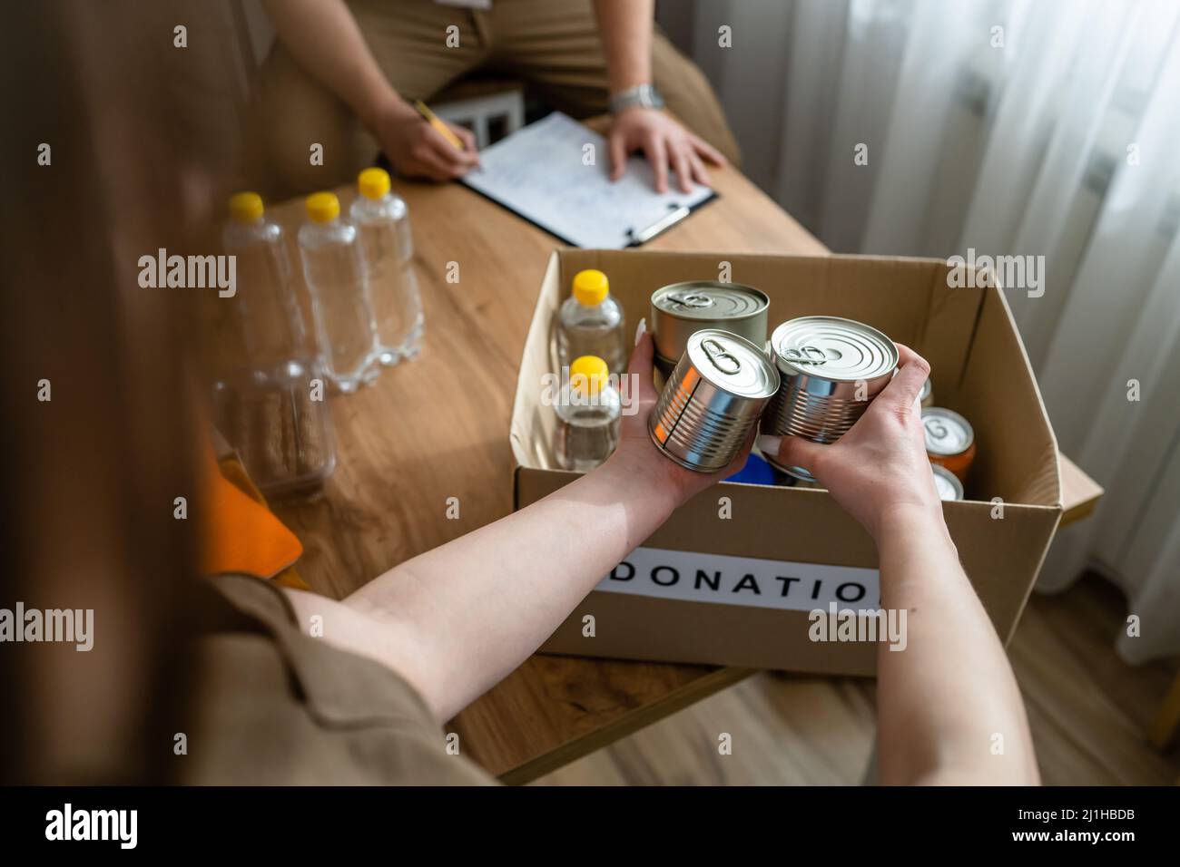 Nahaufnahme von Konserven auf dem Tisch, bereit für die Spende bei der Stiftung der Wohltätigkeitsorganisation, mit unbekannter kaukasischer Frau, die freiwillig Sachen in die Box legt Stockfoto