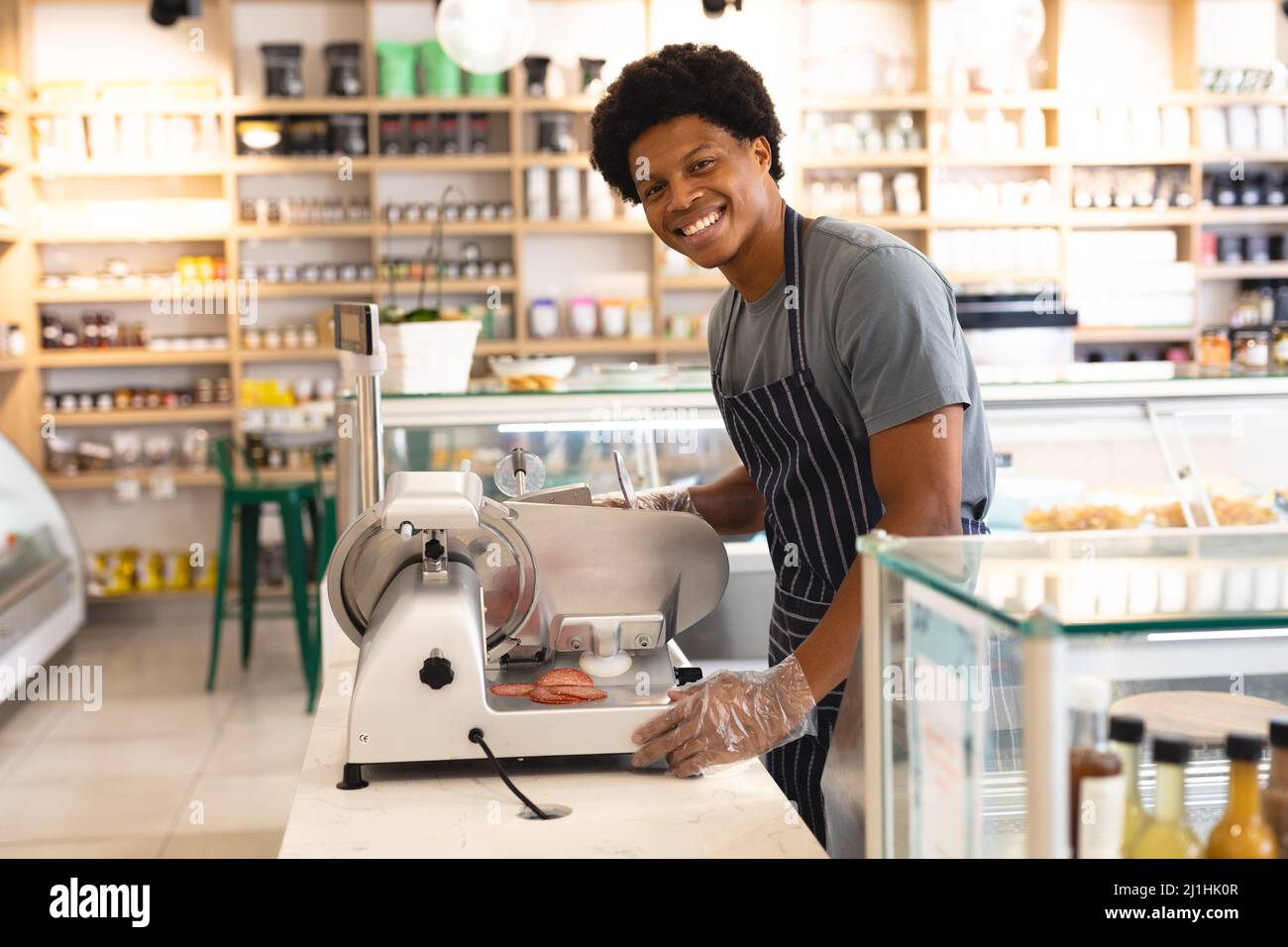 Porträt eines lächelnden jungen männlichen afroamerikanischen Angestellten, der Fleischschneidemaschine an der Theke im Café verwendet. Unverändert, Cafekultur, Menschen und Beruf c Stockfoto