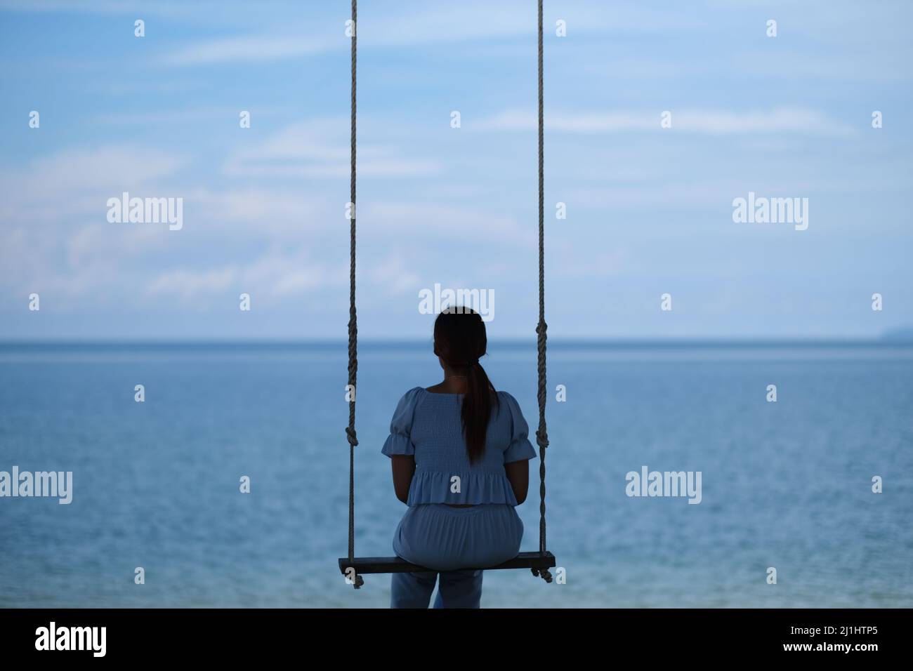 Allein am Meer: Die junge Frau, von hinten betrachtet, entspannt sich auf einer Schaukel mit einer schönen, ruhigen Meereslandschaft vor sich Stockfoto