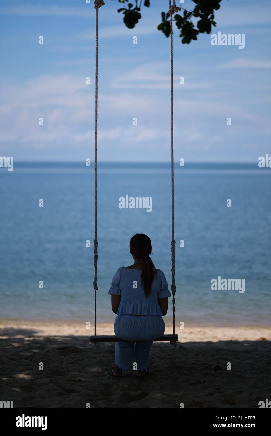 Allein am Meer: Die junge Frau, von hinten betrachtet, entspannt sich auf einer Schaukel mit einer schönen, ruhigen Meereslandschaft vor sich Stockfoto