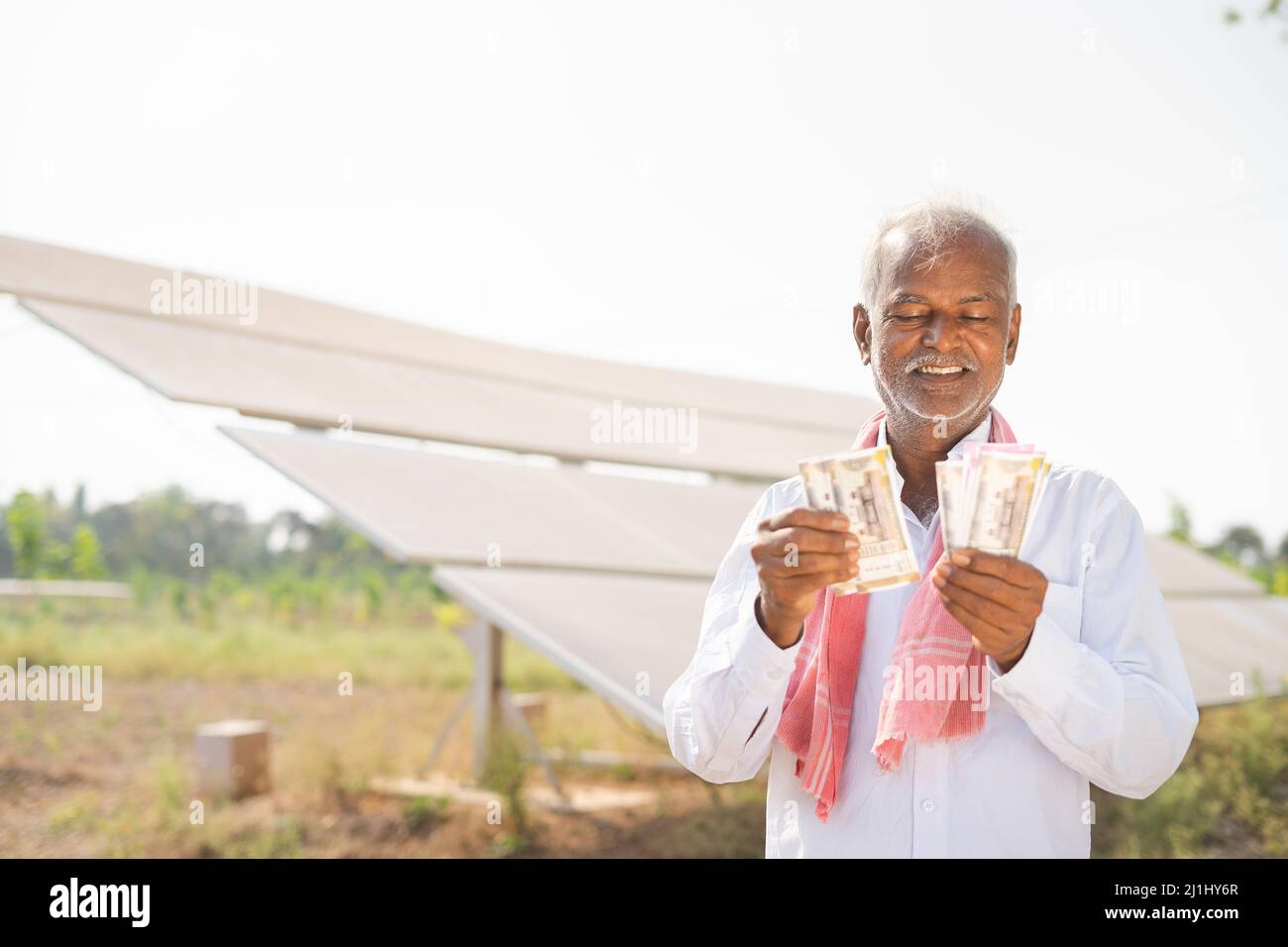 Glücklich lächelnder indischer Landwirt, der Geld couting, während er vor dem Solarpanel auf Ackerland steht - Konzept der Stromeinsparung, Investitionen, Banken Stockfoto
