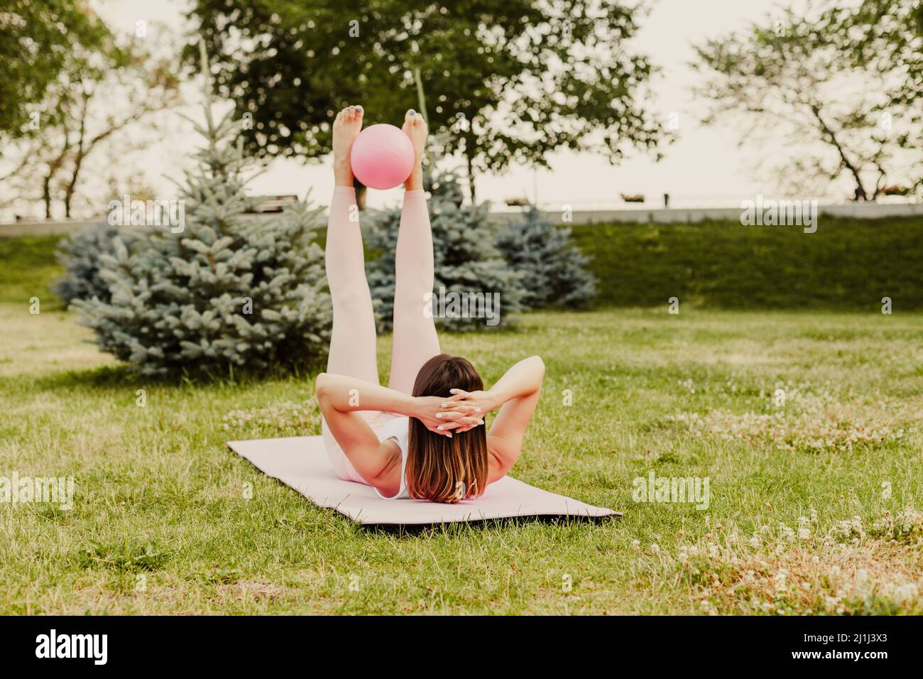 Attraktive kaukasische Frau machen Pilates mit Ball und tun Kreuzungsübungen auf grünem Rasen bei Sonnenuntergang Stockfoto