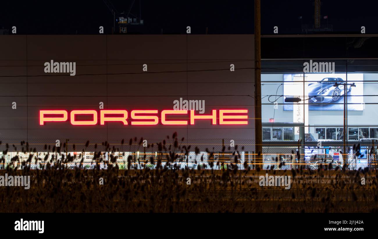 Das Porsche-Logo auf der Seite eines Händlergebäudes ist zu sehen, während es nachts in leuchtendem Rot leuchtet. Stockfoto