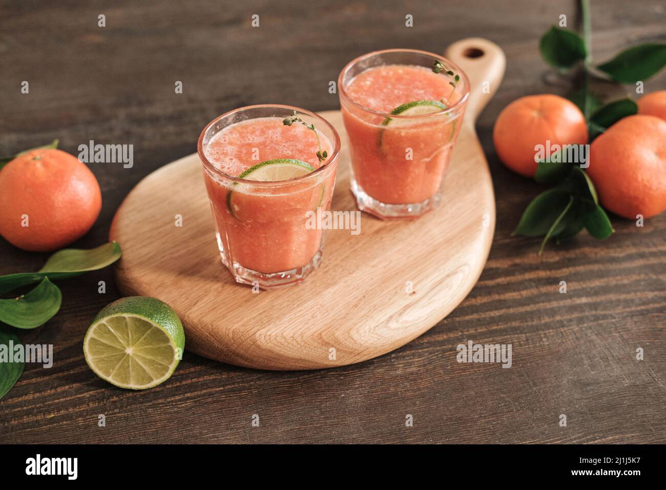 Nahaufnahme eines Holzbretts mit Gläsern frischen Grapefruitsafts, dekoriert mit Limettenscheibe, Mandarinen und Blättern auf einem Holztisch Stockfoto