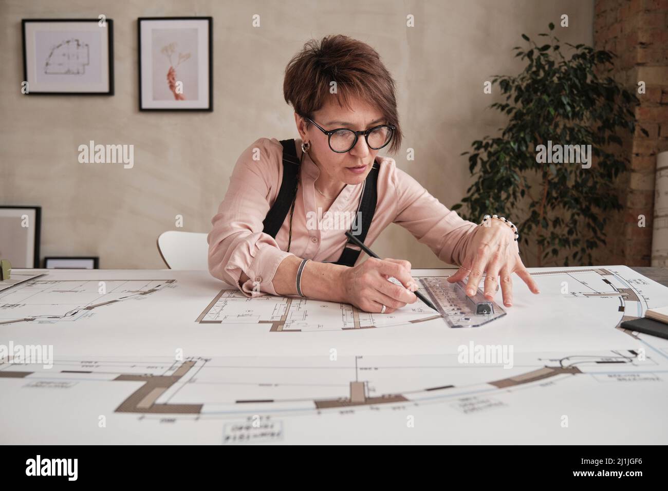 Seriöse reife Frau mit kurzen Haaren sitzt am Tisch im Loft-Büro und mit Lineal, während die Blaupause Zeichnung Stockfoto