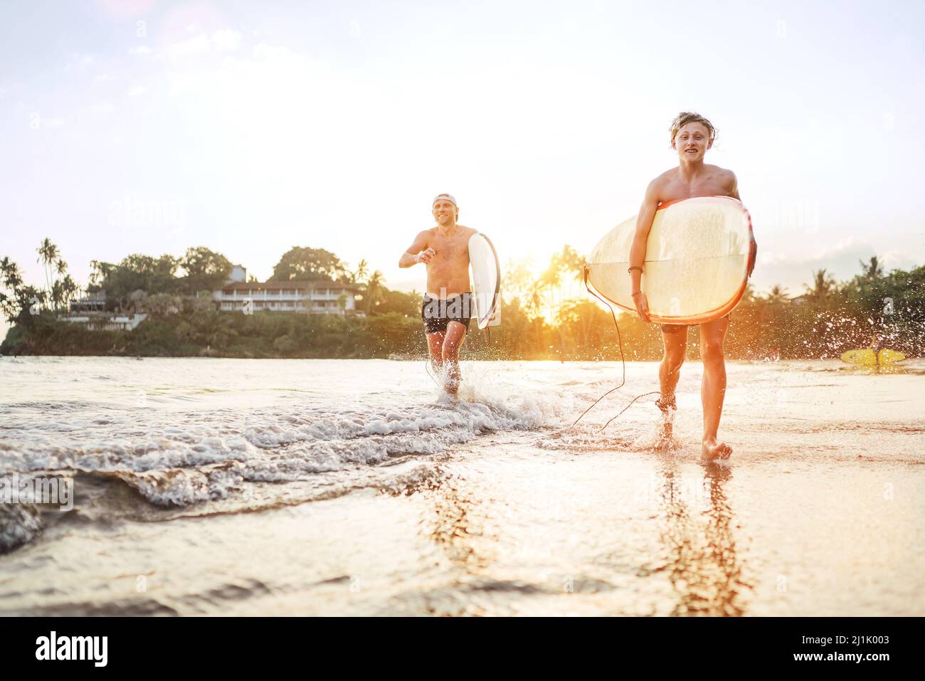 Junger Teenager mit einem Surfbrett, der mit seinem Vater am Sandstrand des Ozeans spazierengeht, nachdem er mit einem wunderschönen Hintergrund bei Sonnenuntergang gesurft hat. Sie sind lächelnd und enjo Stockfoto