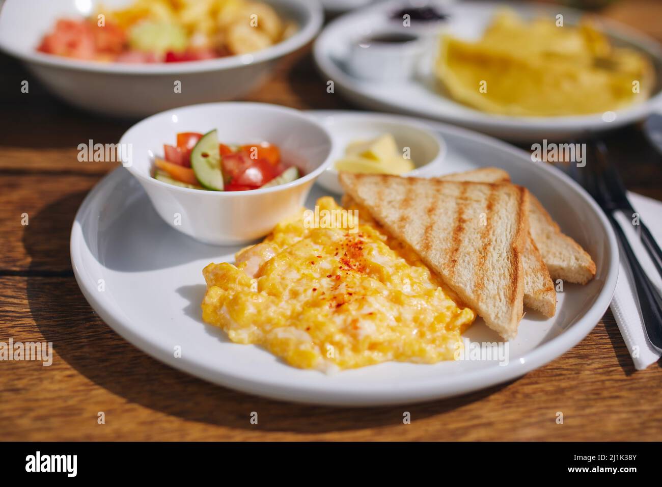 Abwechslungsreiches Frühstück auf Holztisch. Nahaufnahme von Rühreiern, Toast, Brot und Gemüsesalat. Stockfoto