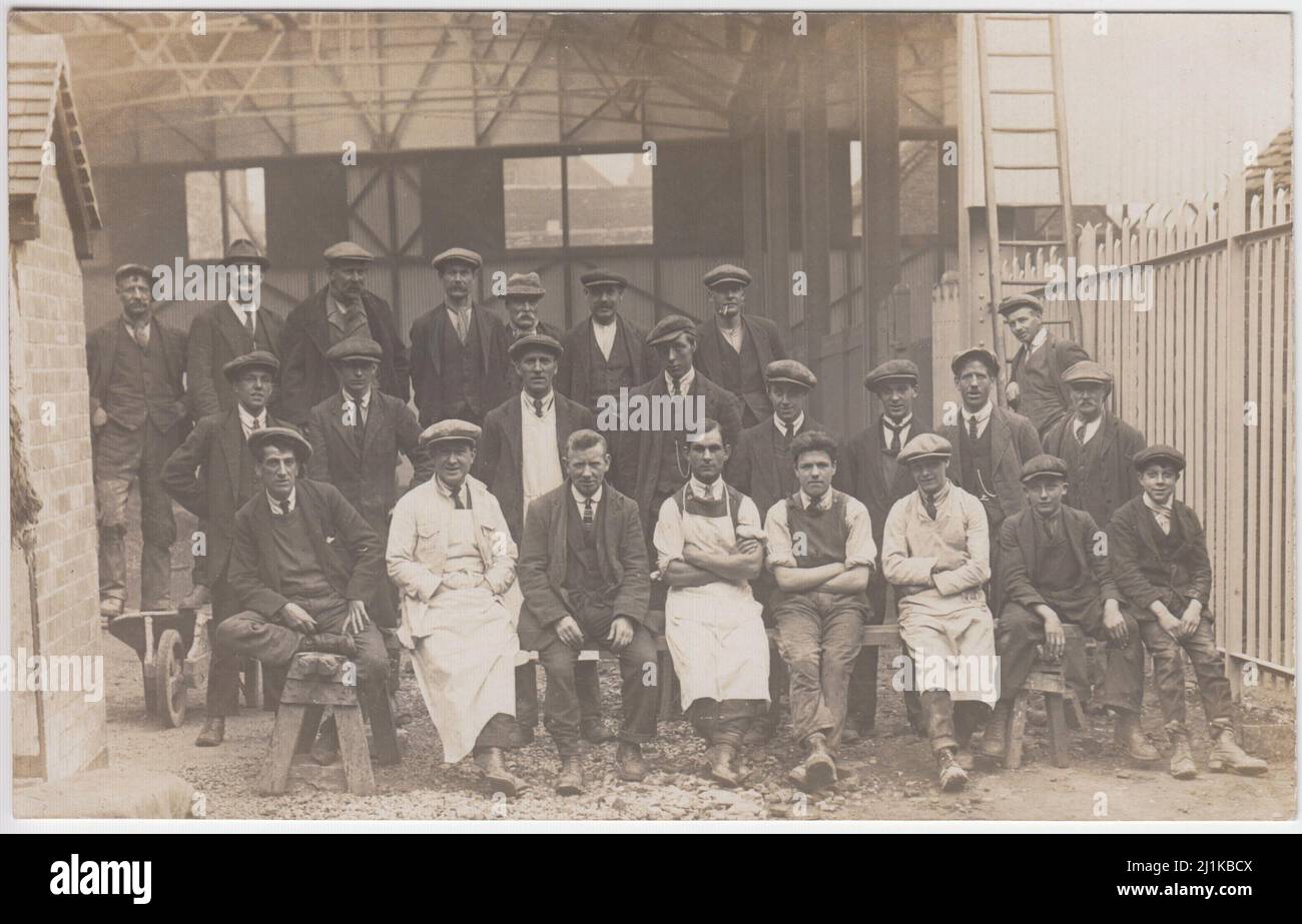 Gruppe von Bauarbeitern, die vor einem Gebäude posieren, Anfang des 20.. Jahrhunderts lehnt sich ein Mann auf eine Leiter, ein Paar steht auf einer Schubkarre und mehrere sitzen auf Werkbänken Stockfoto