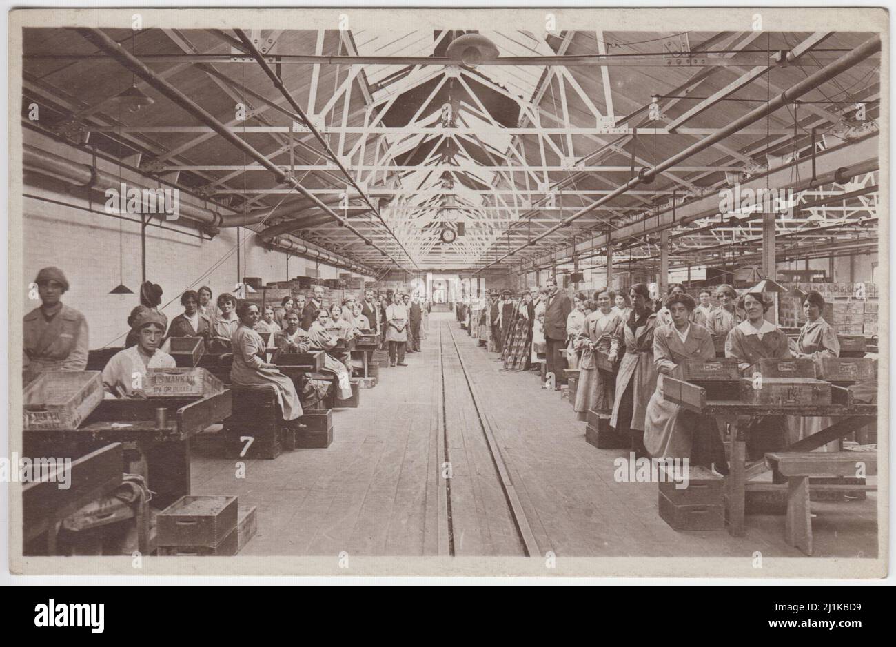 Royal Small Arms Factory, Enfield, Hauptmaschinenfabrik, erster Weltkrieg: Munitionsarbeiterinnen, die auf Werkbänken sitzen, Munitionskisten (darunter eine mit der Aufschrift „US Mark VII 174 Gr. Bullet') auf den Bänken. Männliche Arbeiter stehen neben Kisten auf der Rückseite der Werkstatt. Fotopostkarte, verkauft von Wakefield's, Ealing, London. Stockfoto