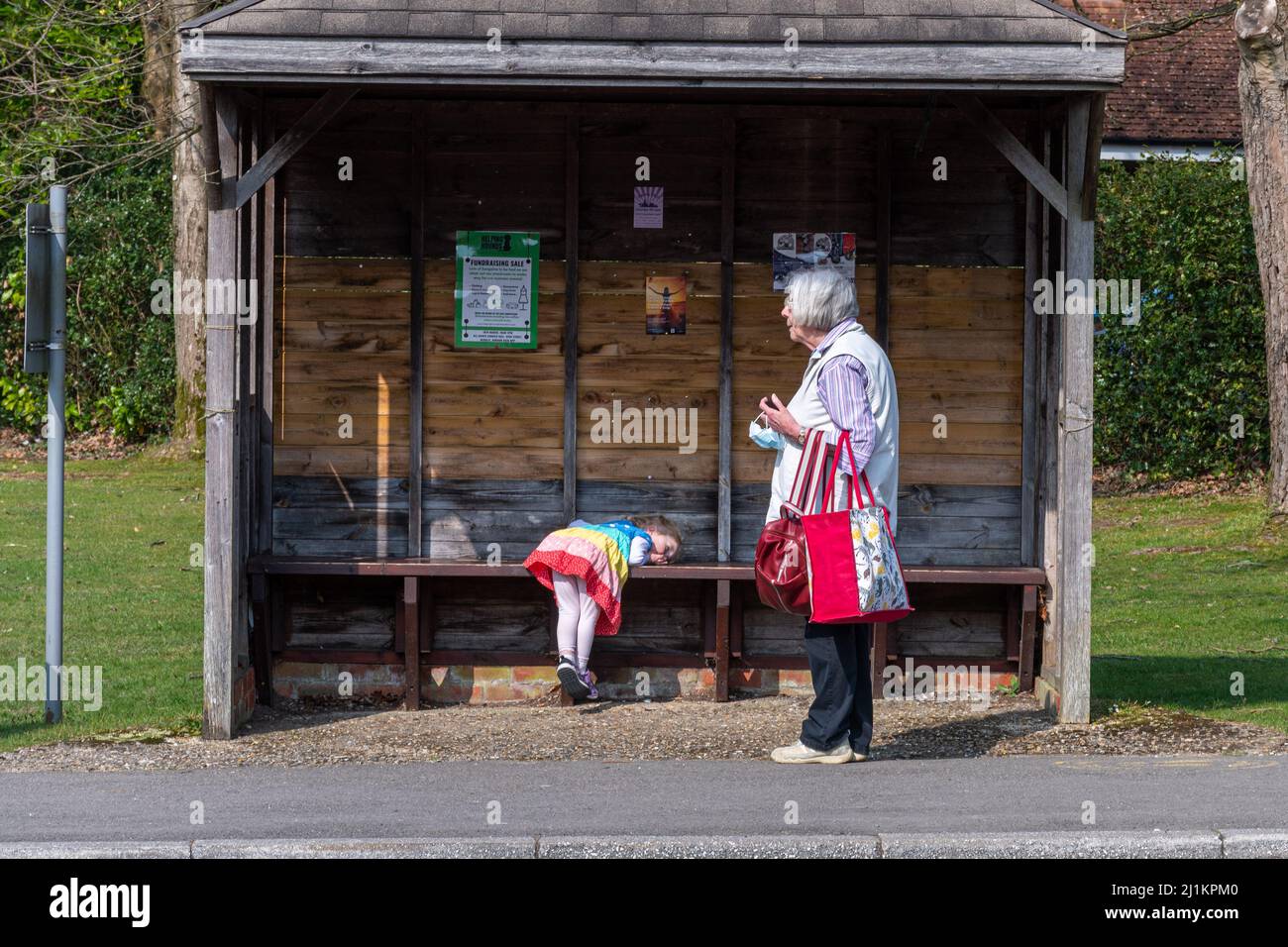 Großmutter, die sich um ihre junge Enkelin gefreut hat, die an einer Bushaltestelle in England, Großbritannien, wartet. Das kleine Mädchen sieht gelangweilt oder müde aus. Stockfoto