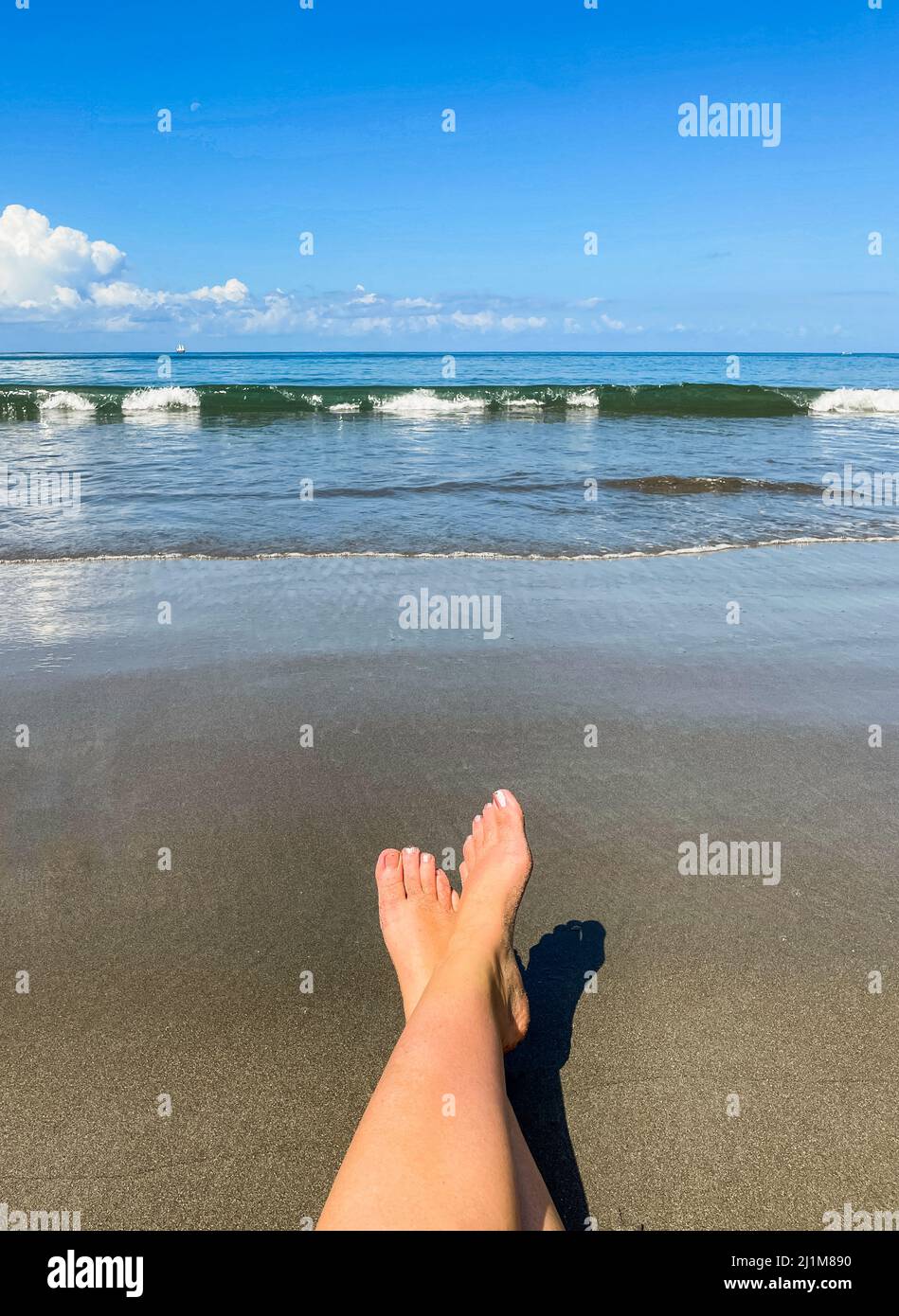 Die ausgestreckten Füße der Frau entspannen sich am Strand gegen das Meer. Stockfoto