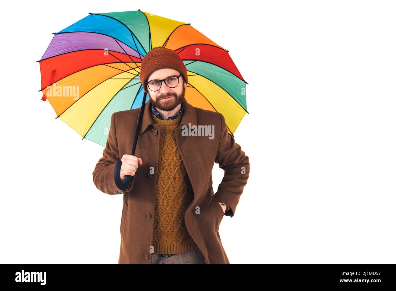 Porträt eines jungen gut aussehenden kaukasischen Mann in Brille und Hut hält einen bunten Regenschirm, hält eine andere Hand in der Tasche und Blick auf die Kamera Positivität Optimismus Wetter Konzept braun casual Kleidung isoliert weißen Hintergrund Studio-Aufnahme . Hochwertige Fotos Stockfoto