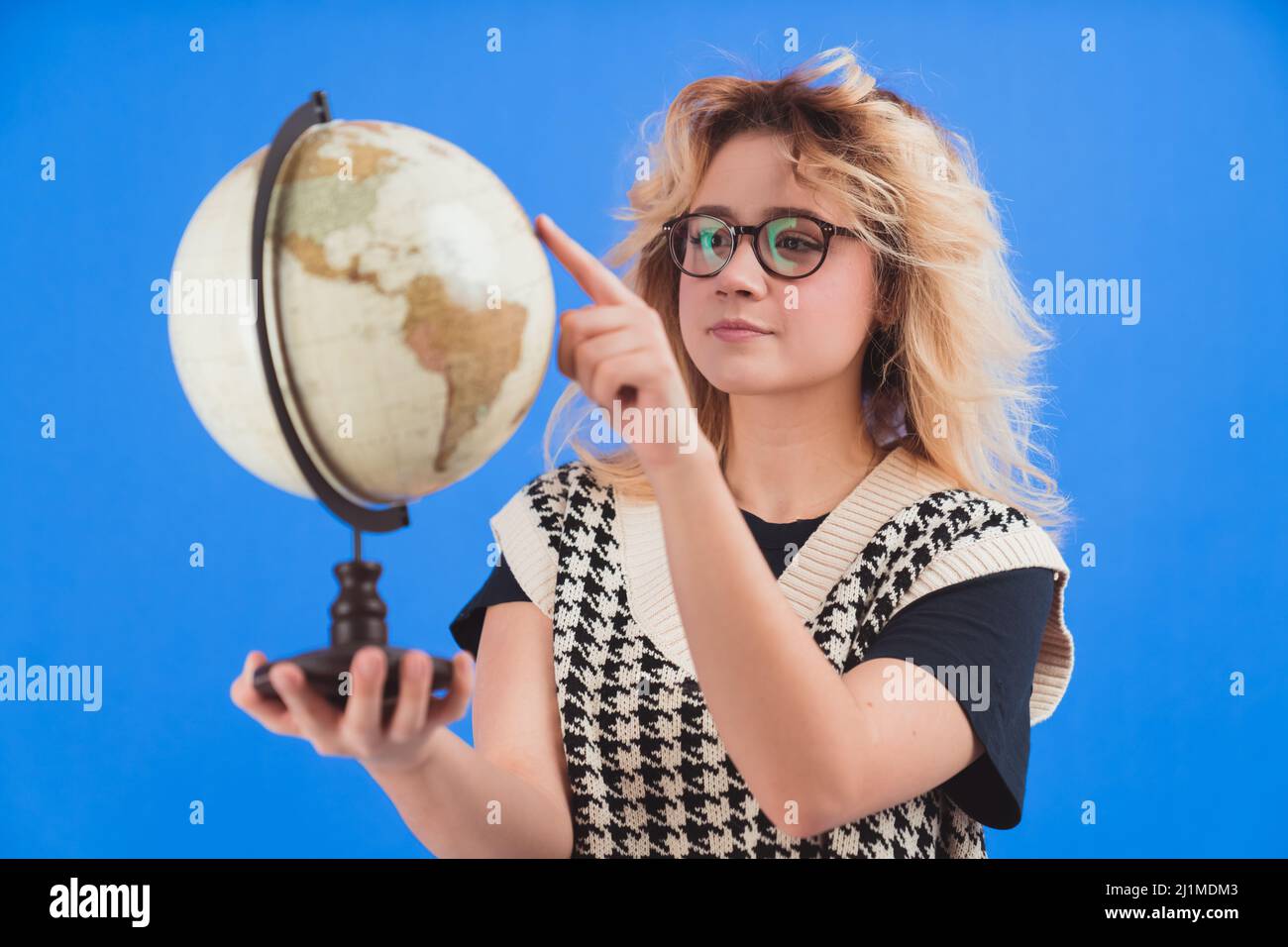 Das junge kaukasische blonde Mädchen mit Brille hält einen künstlichen Globus und zeigt mit ihrem Zeigefinger auf den blauen Hintergrund. Isolierte Studioaufnahme. . Hochwertige Fotos Stockfoto