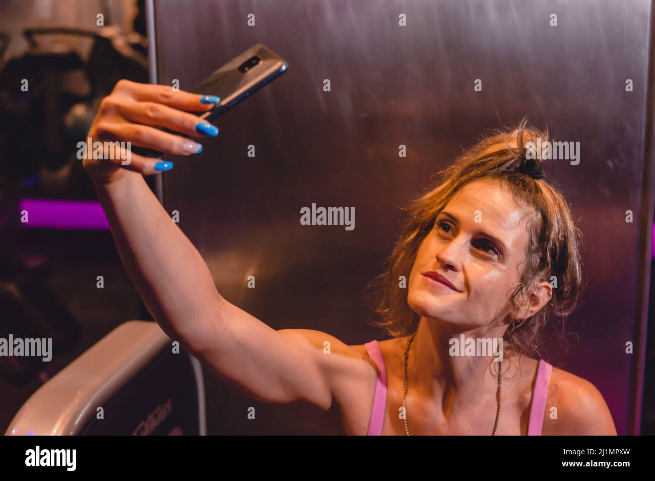 Die schöne junge kaukasische blonde lateinerin aus argentinien im Fitnessstudio macht lächelnd ein Selfie mit ihrem Handy. Stockfoto