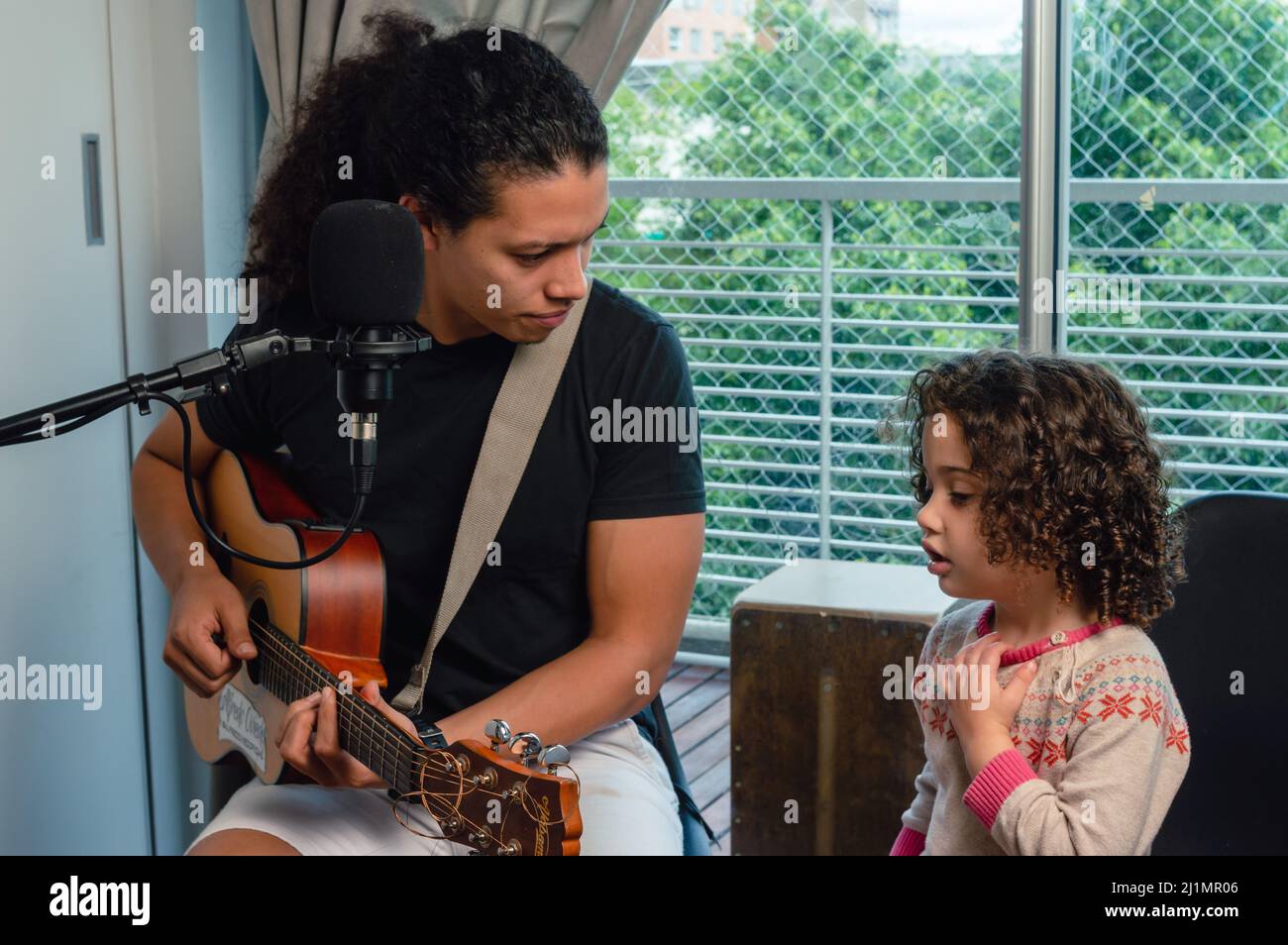 Der kaukasische junge Vater spielt Gitarre und sieht seiner Tochter zu, die mit ihm singt, drinnen im heimischen Studio sitzt und in der Familie zusammen Musik macht. Stockfoto