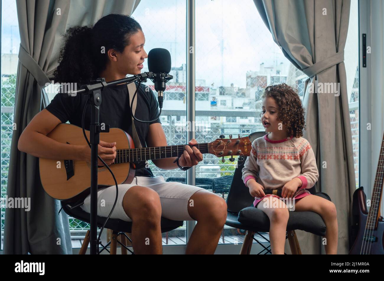 Der junge lateinamerikanische venezolanische Vater singt mit der Gitarre zu seiner Tochter, die in ihrem heimischen Musikstudio sitzt und gemeinsam Musik zu Hause macht, Musik und Stockfoto
