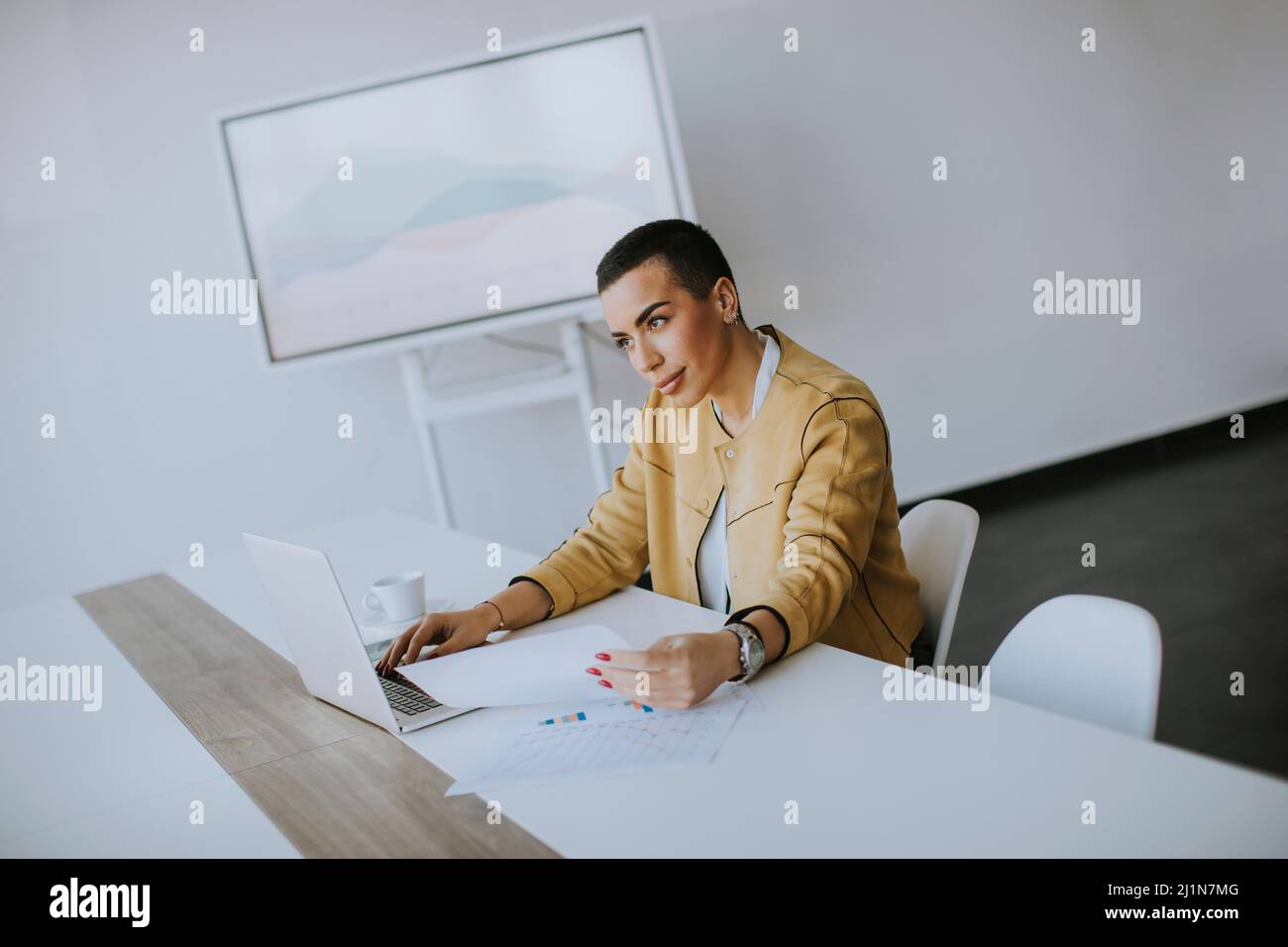 Junge Frau mit kurzen Haaren, die am Laptop arbeitet, während sie im Büro sitzt Stockfoto