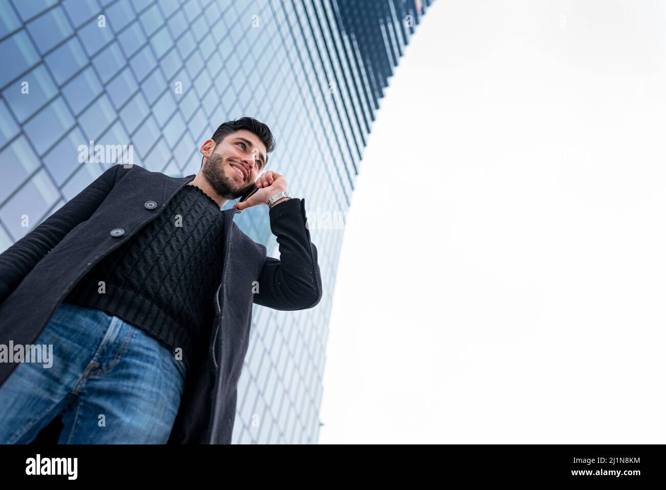 Selbstbewusster und erfolgreicher junger Geschäftsmann, der mit einem Smartphone telefoniert, verwackelte finanzielle Wolkenkratzer im Hintergrund, Unternehmer im Fernarbeiten, glücklich Stockfoto