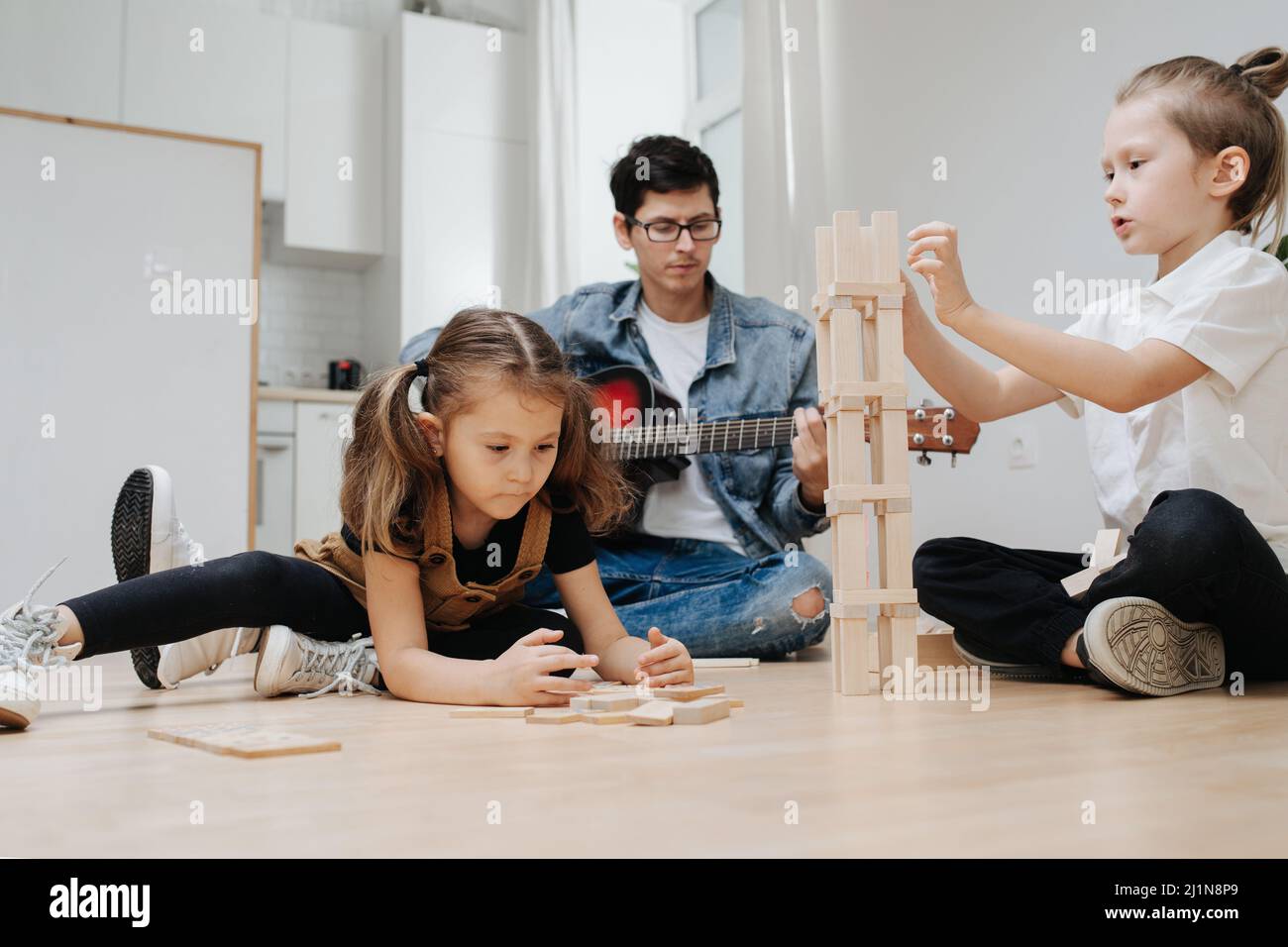 Junge spielt auf dem Küchenboden mit Holzblöcken neben seiner Schwester, die den Turm baut. Fröhlicher Vater spielt Gitarre im Hintergrund. Stockfoto