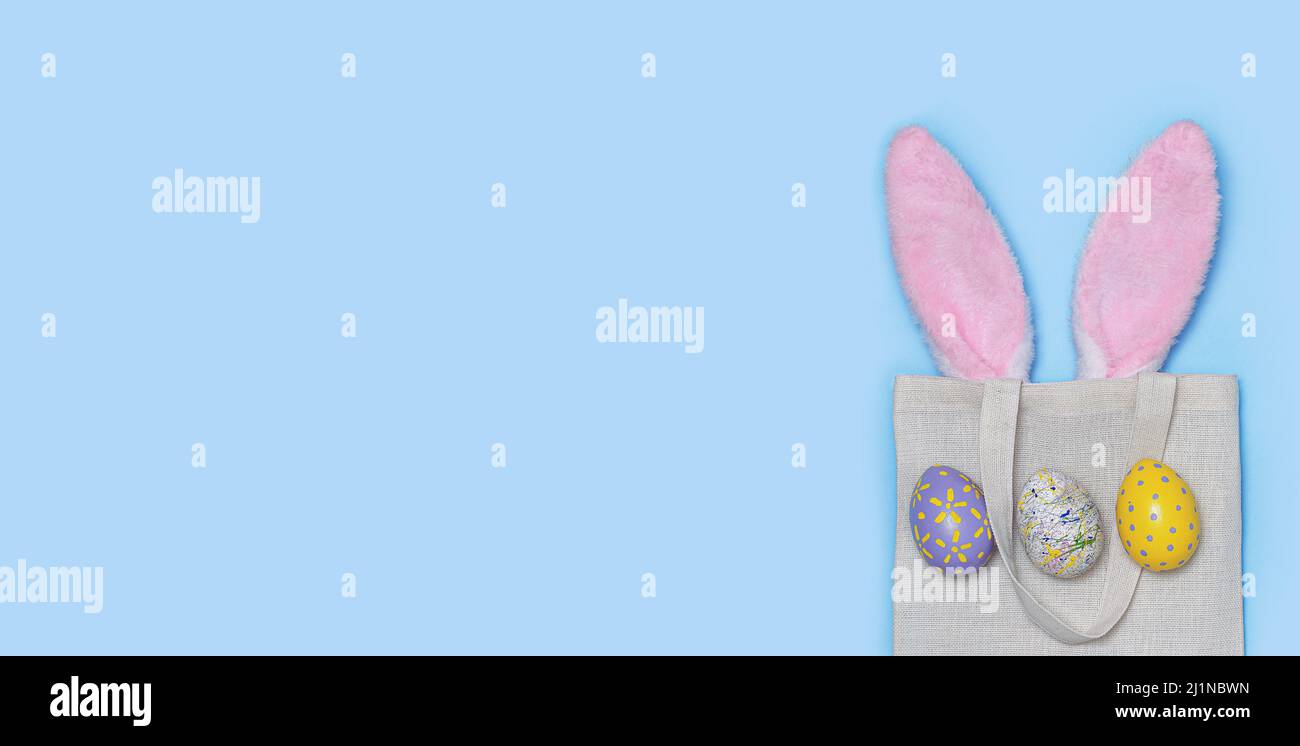 Banner. Rosafarbene Osterhasen-Ohren ragen aus dem wiederverwendbaren Stoffbeutel mit Painted Eggs auf blauem Hintergrund. Happy Easter Minimales Konzept. Ein Osterfest ca. Stockfoto