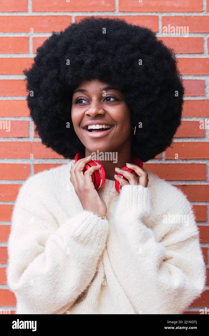 Porträt einer fröhlichen afroamerikanischen Frau mit weißem, perfektem Lächeln, Afro-Frisur, weißem Pullover und roten Kopfhörern im Freien, während sie wegschaut Stockfoto