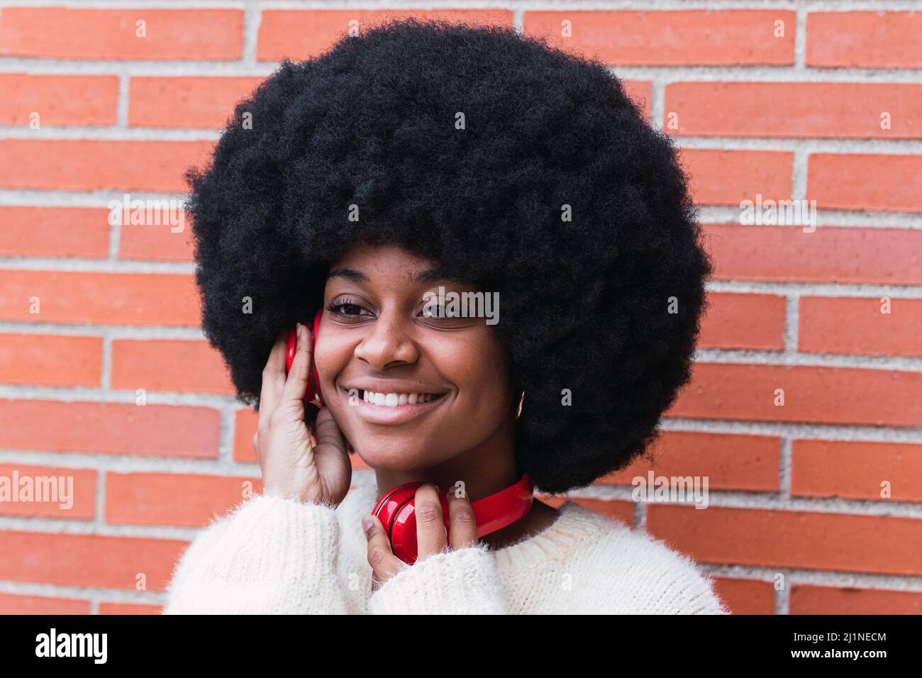 Seitenansicht Porträt einer fröhlichen schwarzen Frau mit weißem perfekten Lächeln, Afro-Frisur, weißem Pullover und roten Kopfhörern im Freien. Zahnarztkonzept Stockfoto