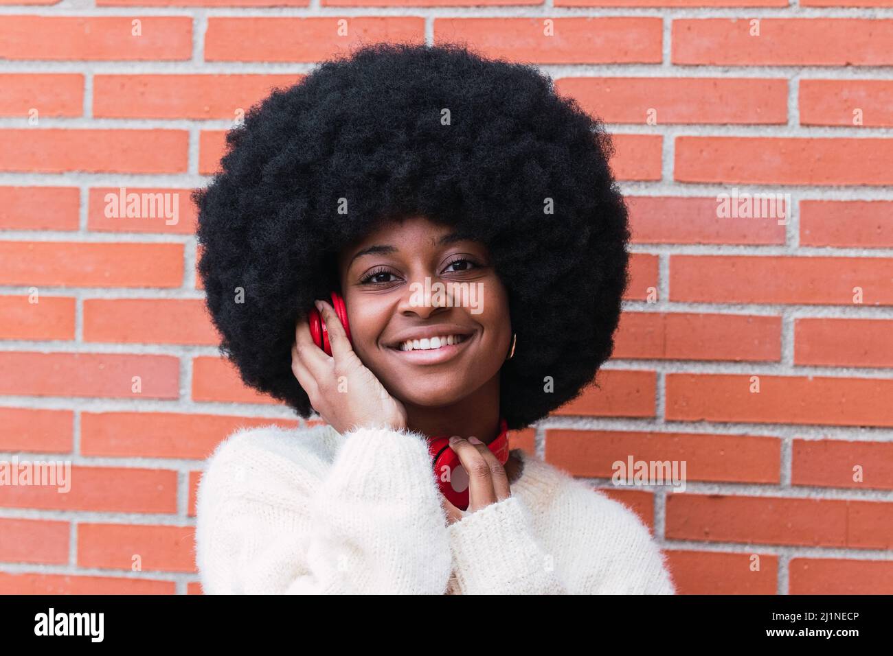 Porträt einer fröhlichen afroamerikanischen Frau mit weißem, perfektem Lächeln, Afro-Frisur, weißem Pullover und roten Kopfhörern im Freien, während sie auf die Kamera schaut Stockfoto