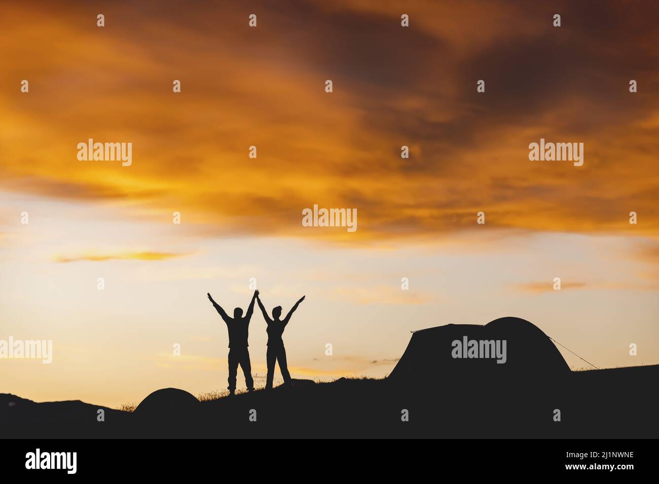 Silhouette eines Paares mit erhöhten Armen und Zelt auf dem Hintergrund des stimmungsvollen Sonnenuntergangs Himmel Stockfoto