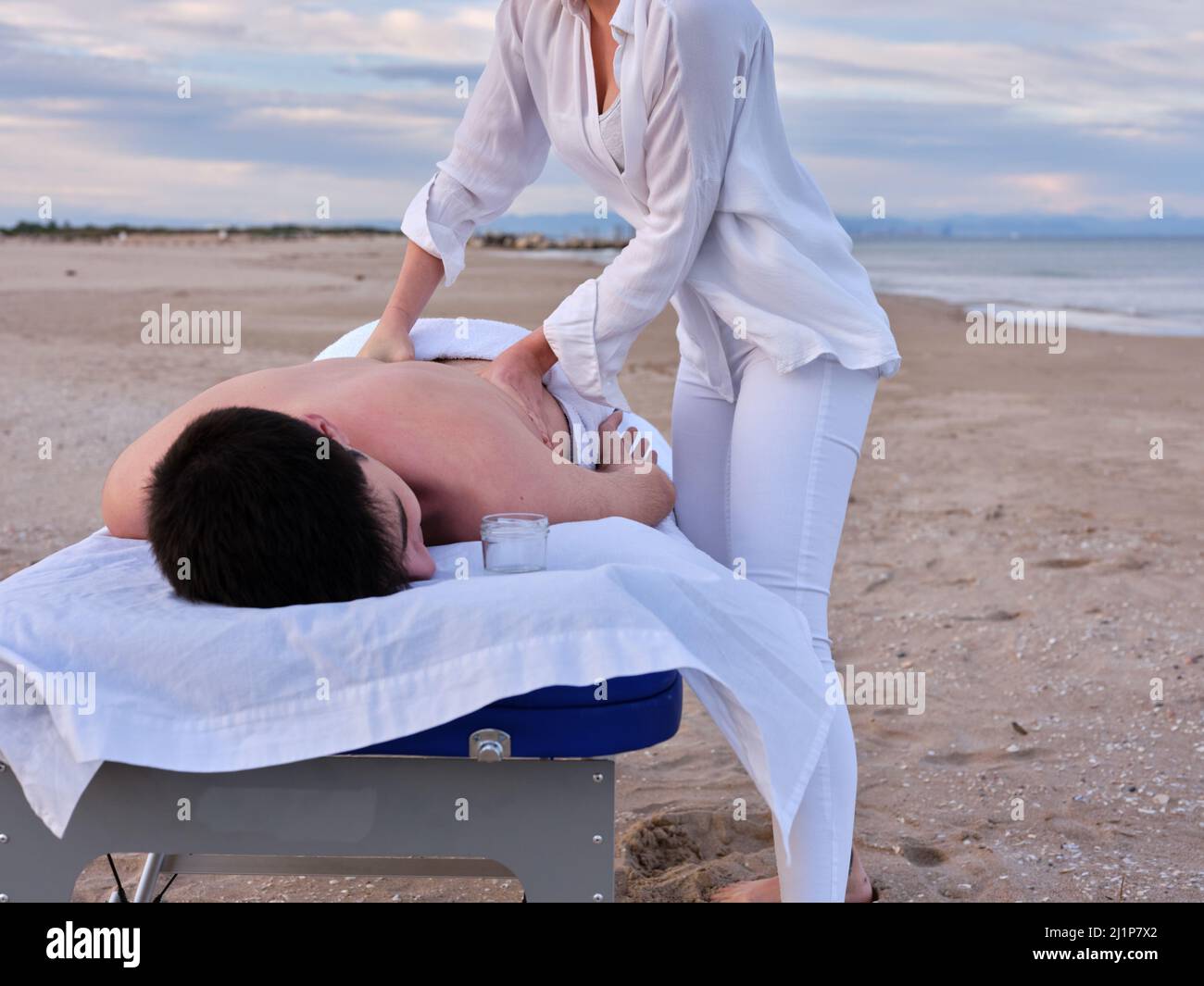 Ein junger Mann, der auf einem Massageliegen liegt und eine Chiromassage am Strand von Valencia von einem nicht erkennbaren jungen Chiromassage-Therapeuten erhält. Stockfoto