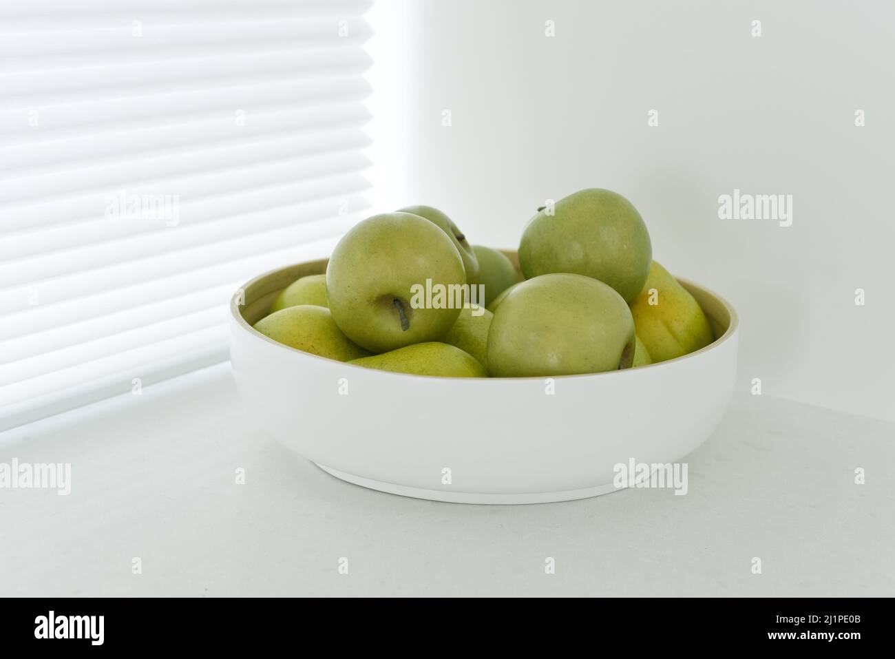 Eine weiße Schale mit grünen Äpfeln von Granny Smith sitzt auf einer weißen Küchentheke vor den Fensterrollos mit Lattenrost Stockfoto
