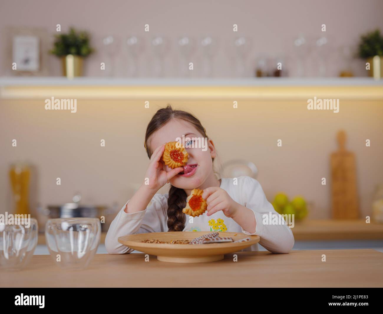 Glückliches kleines Mädchen lächelt glücklich, wenn sie Kekse isst, die zu Hause am Tisch in der Küche sitzen. Lustiges Baby jagt nach leckeren Cookies Stockfoto