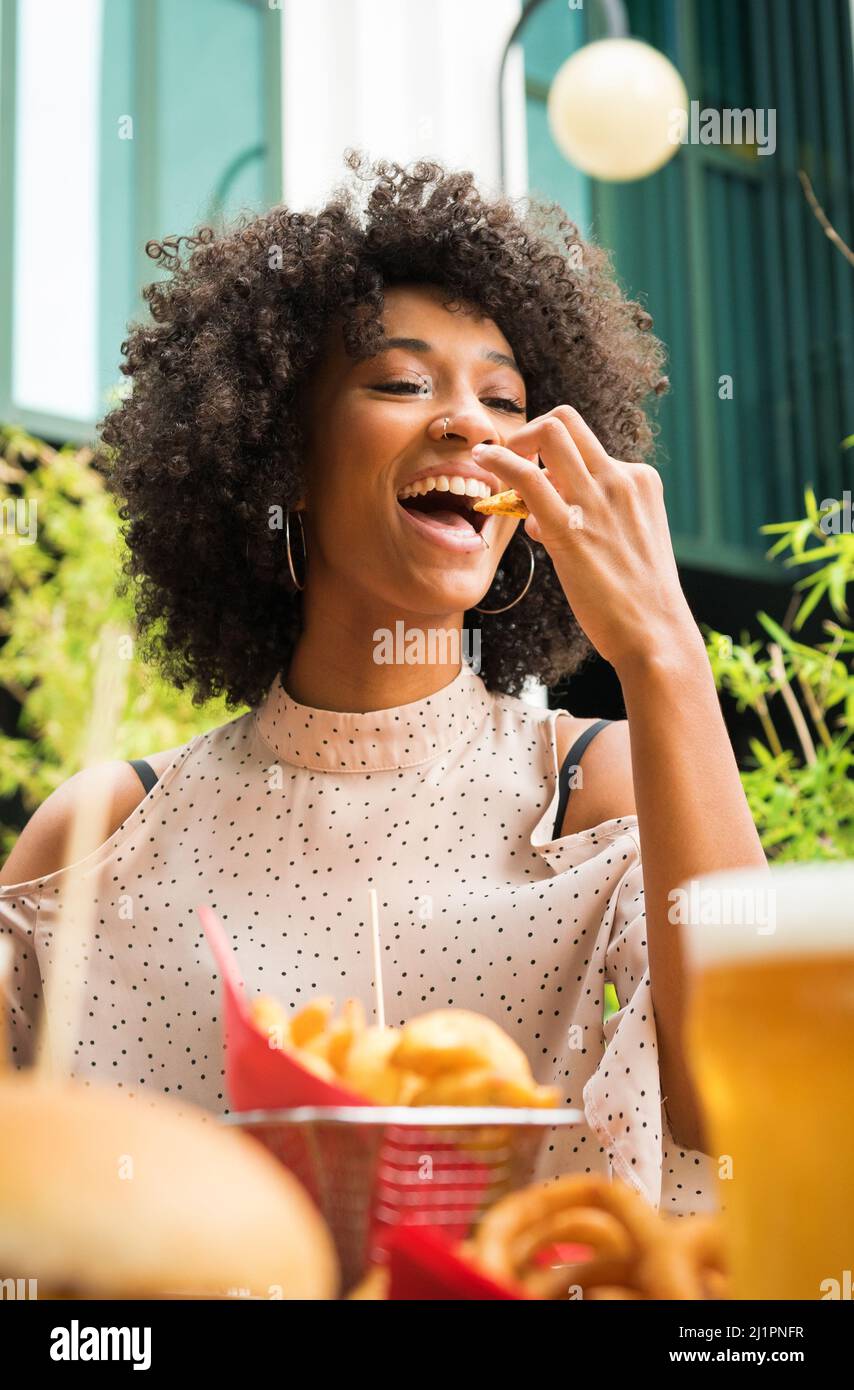 Lächelnd schöne junge glückliche schwarze Frau mit Nasenpiercing, die Kartoffelkeile in einem Pub in einem niedrigen Winkel über Essen und Gläser Bier isst Stockfoto
