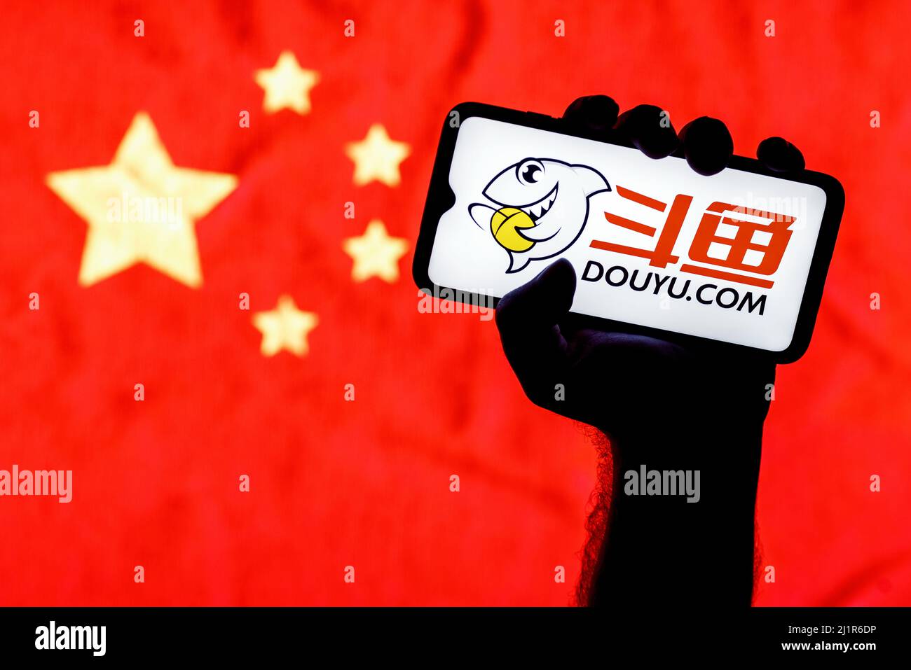 Ein Smartphone mit dem Logo des DouYu Live Streaming Service auf dem Bildschirm in einer zusammengeballten Hand auf dem Hintergrund einer chinesischen Flagge. Stockfoto