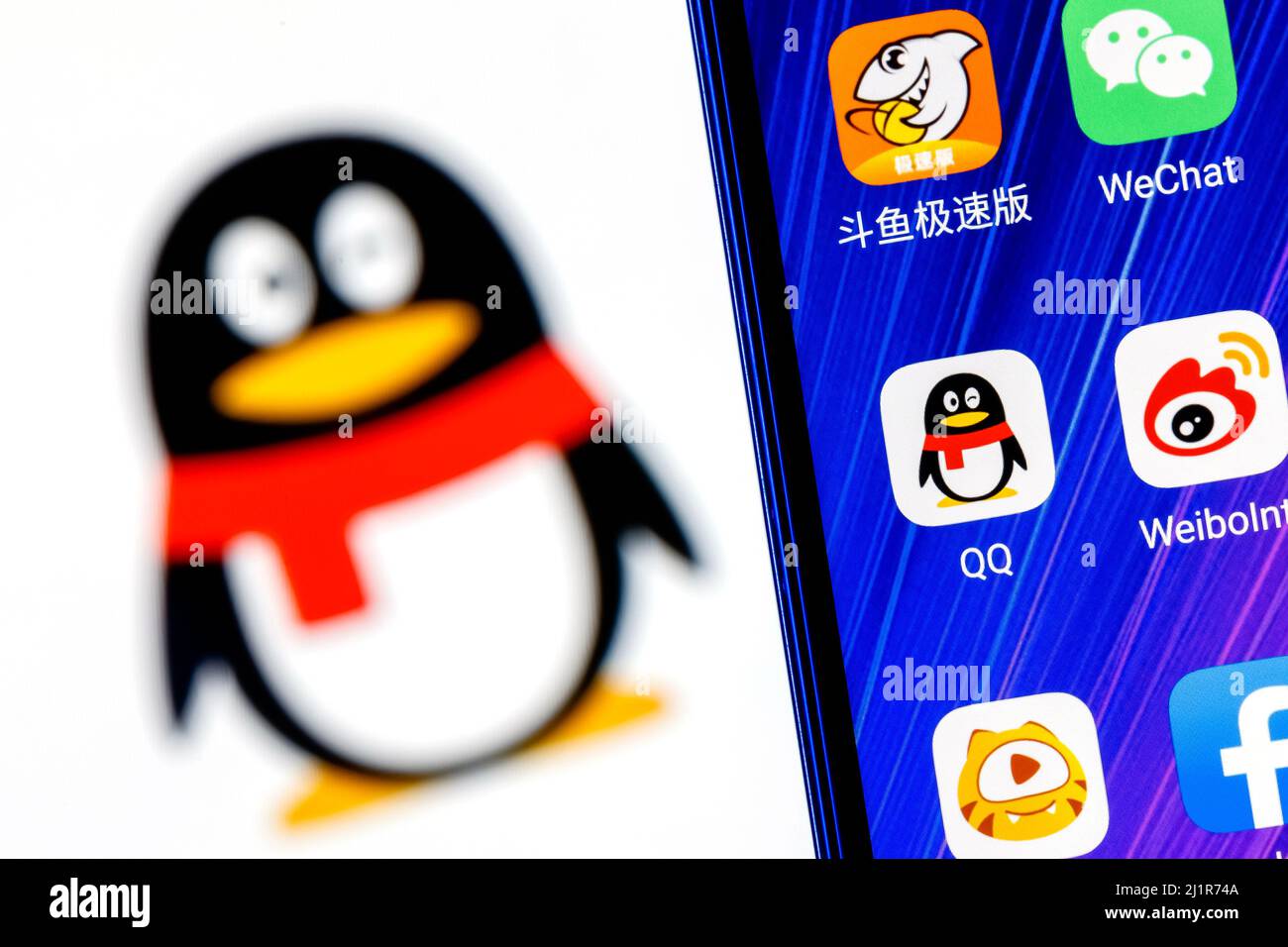 Das Symbol der chinesischen Nachrichtendienst-Anwendung Tencent QQ unter anderem auf dem Smartphone-Bildschirm. Auf dem Hintergrund ist das QQ-Logo zu sehen. Stockfoto