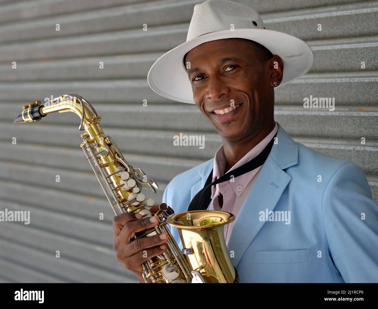 Schöner eleganter junger afro-kubanischer Saxophonist mit weißem Panamahut hält sein glänzend poliertes Saxophon und lächelt für den Zuschauer. Stockfoto