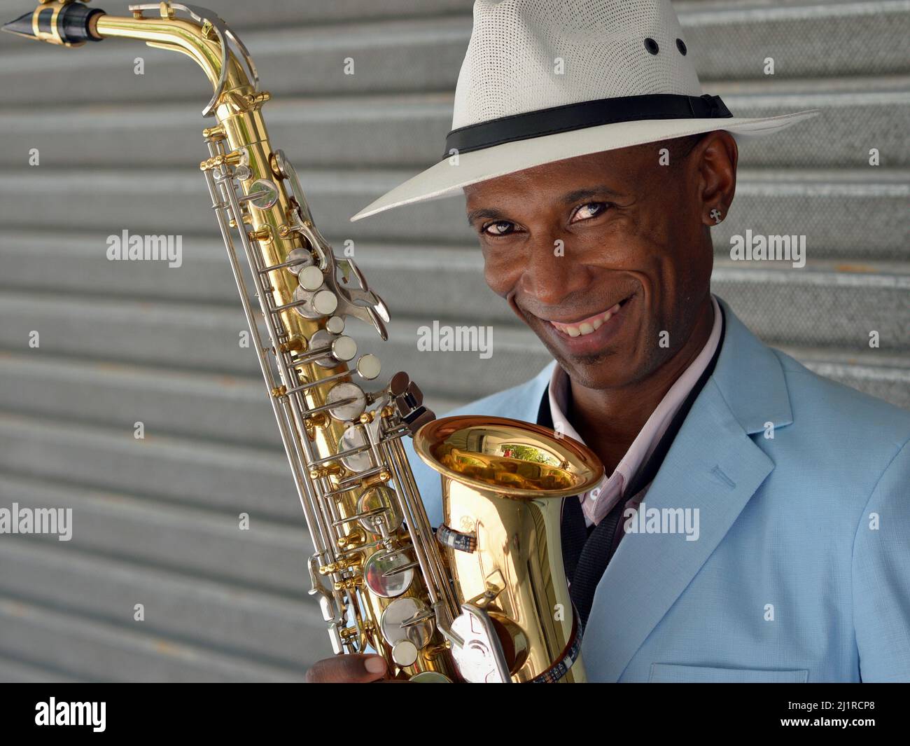 Schöner eleganter junger afro-kubanischer Saxophonist mit weißem Panamahut hält sein glänzend poliertes Saxophon und lächelt für den Zuschauer. Stockfoto