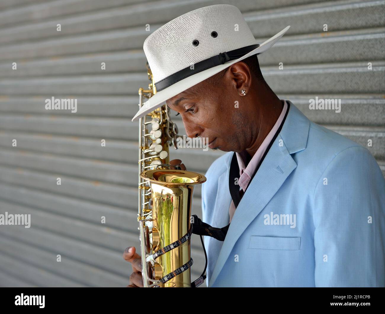 Der hübsche, elegante junge, lustige afro-kubanische Saxophonist trägt einen weißen panamahut und blickt in sein Saxophon hinab. Stockfoto
