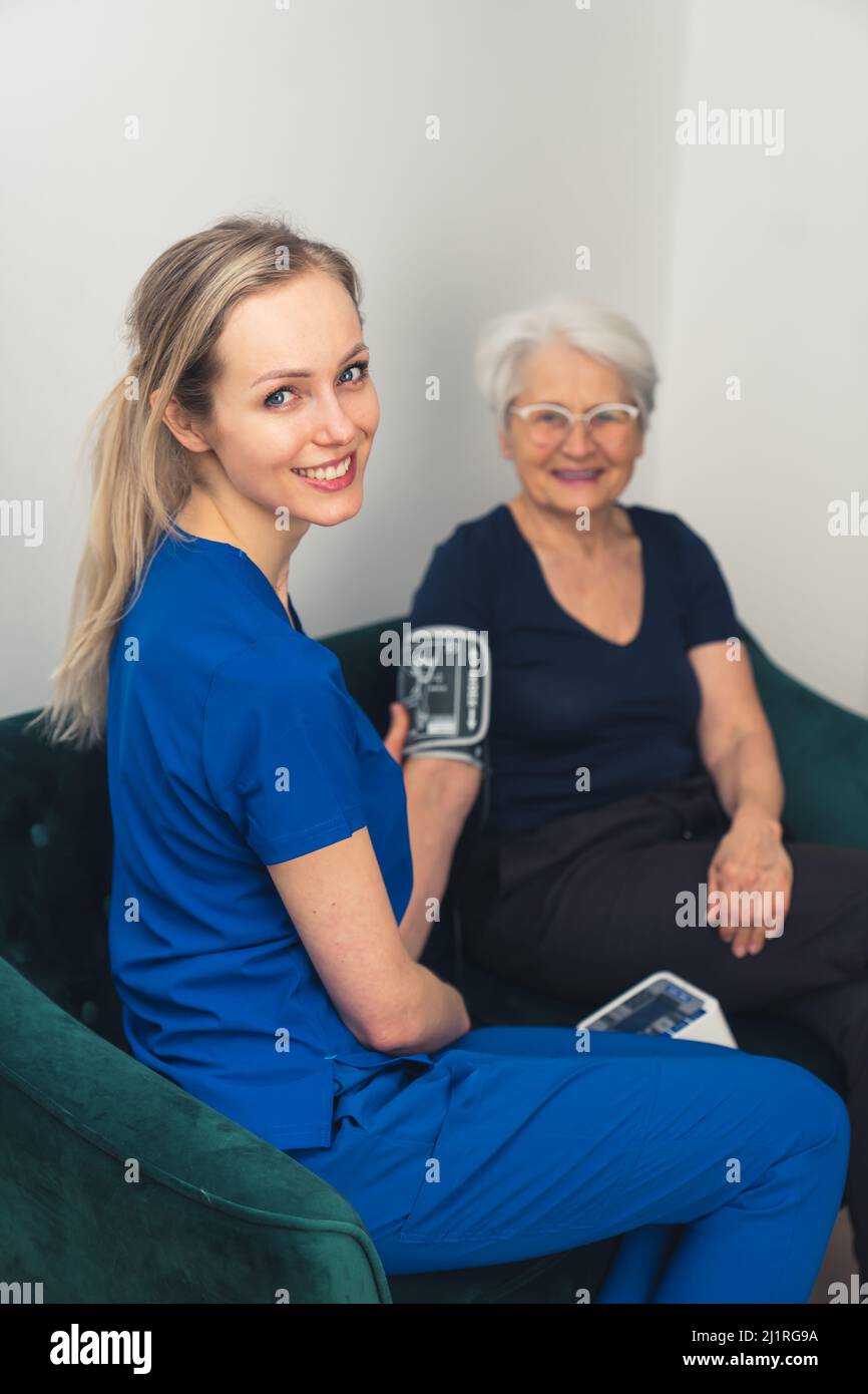 Sich um das eigene Gesundheitskonzept kümmern. Die ältere Oma und ihre schöne junge Pflegekraft sitzen während ihres Hausbesuches auf einem Sofa zusammen. Hochwertige Fotos Stockfoto