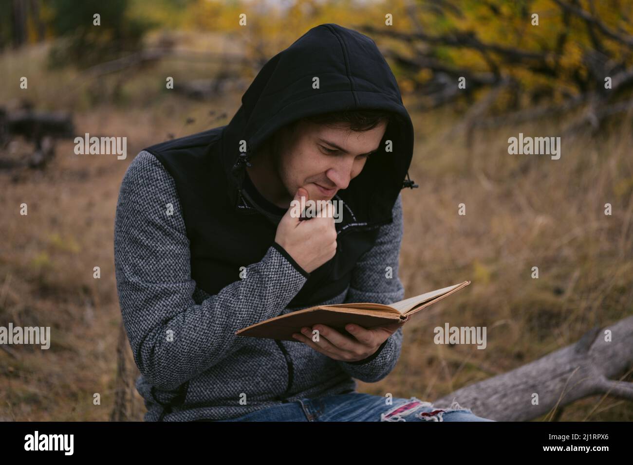 Ein junger, gutaussehender Mann in lässiger Kleidung sitzt auf einem Holzbuch und liest im Herbstwald ein Buch Stockfoto